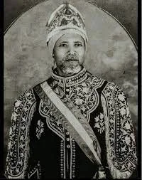 Black and white portrait of a man in traditional regal attire, wearing a crown and ornate jewelry.