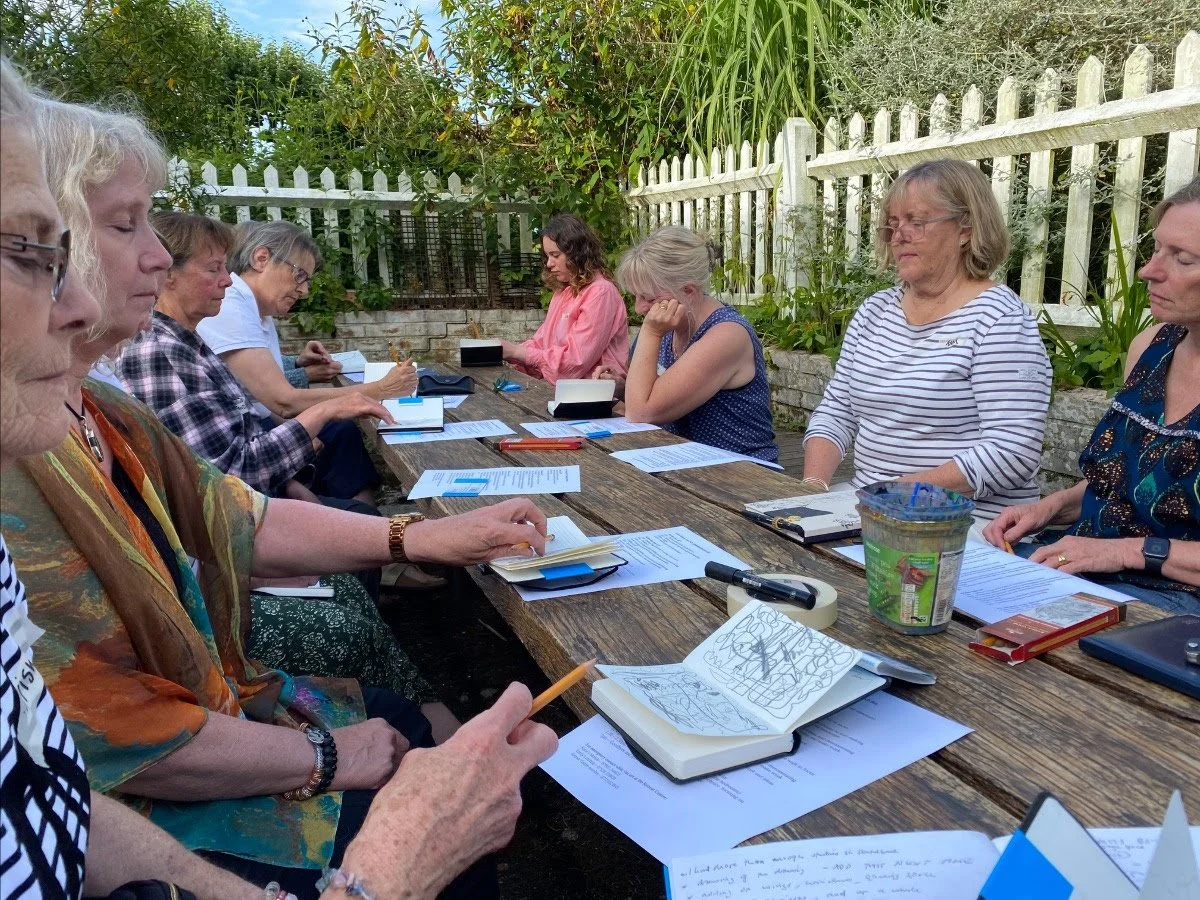 A group of women sitting at a long outdoor wooden table, engaged in a meeting or discussion, with papers, notebooks, and writing utensils in front of them, surrounded by a white picket fence and lush greenery.