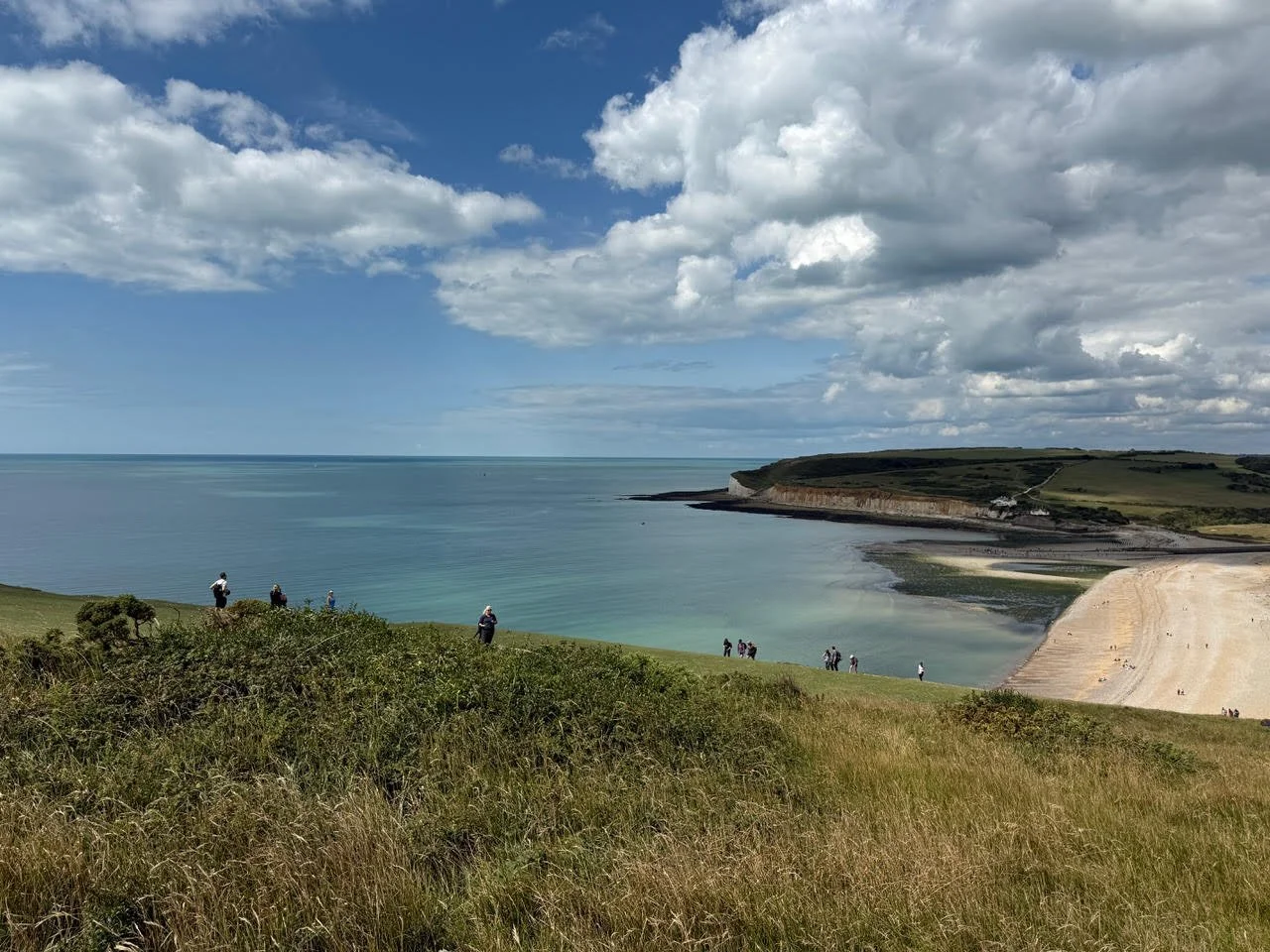 A scenic coastal landscape with grassy hills, a sandy beach, calm ocean waters, and a partly cloudy sky with people walking and enjoying the view.