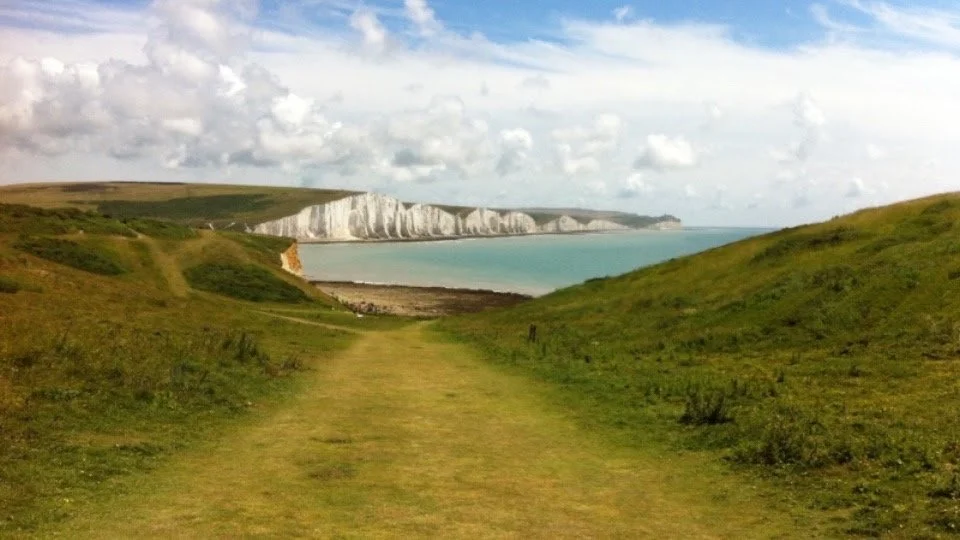 A grassy path leading towards a coastline with white chalk cliffs and a blue ocean under a partly cloudy sky.