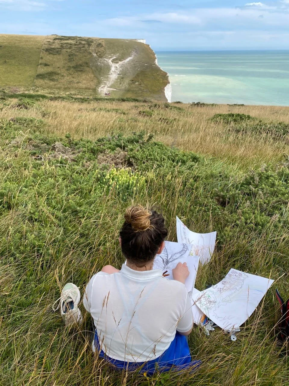 A girl sitting on grass drawing pictures on paper with a scenic view of cliffs, grassy fields, and the ocean in the background.
