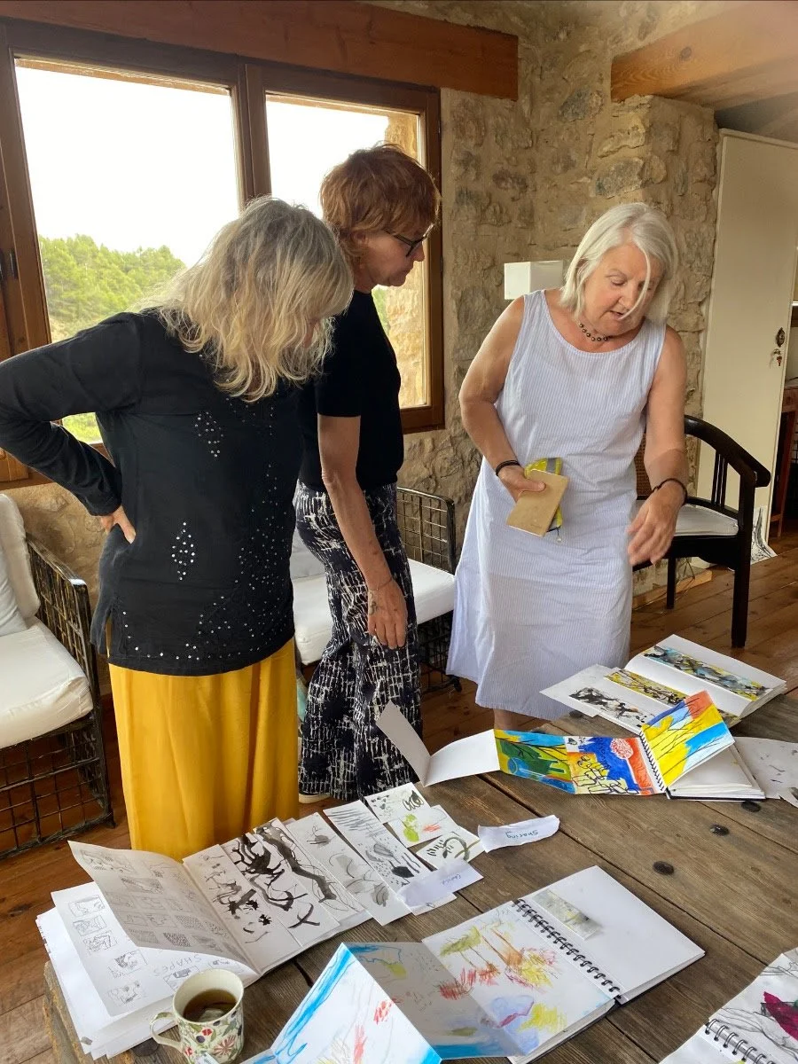 Three women are standing around a wooden table looking at colorful art projects and sketchbooks in a rustic room with stone walls and large windows.