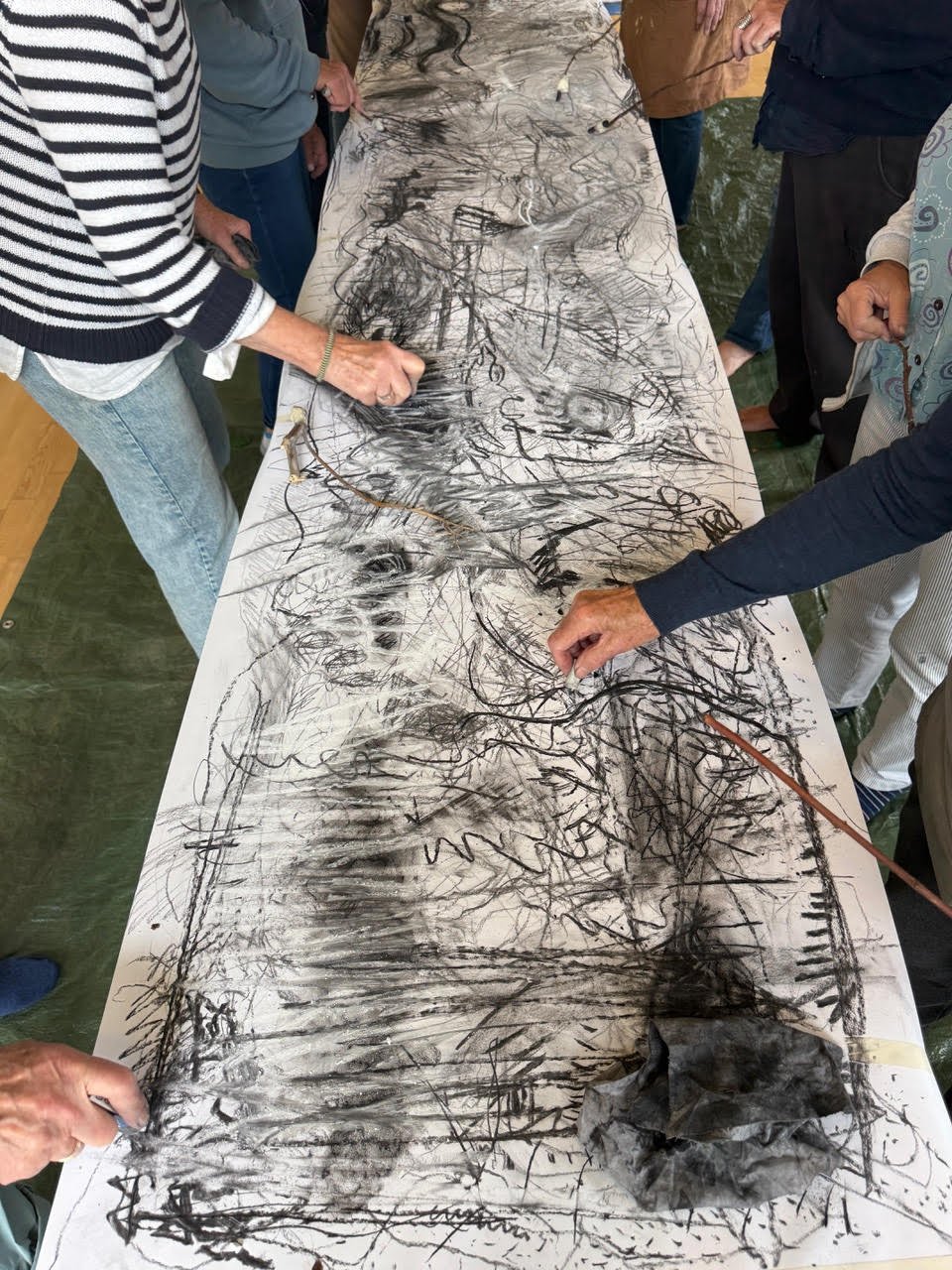 Group of people drawing abstract art with black charcoal on a large white paper spread on a table.