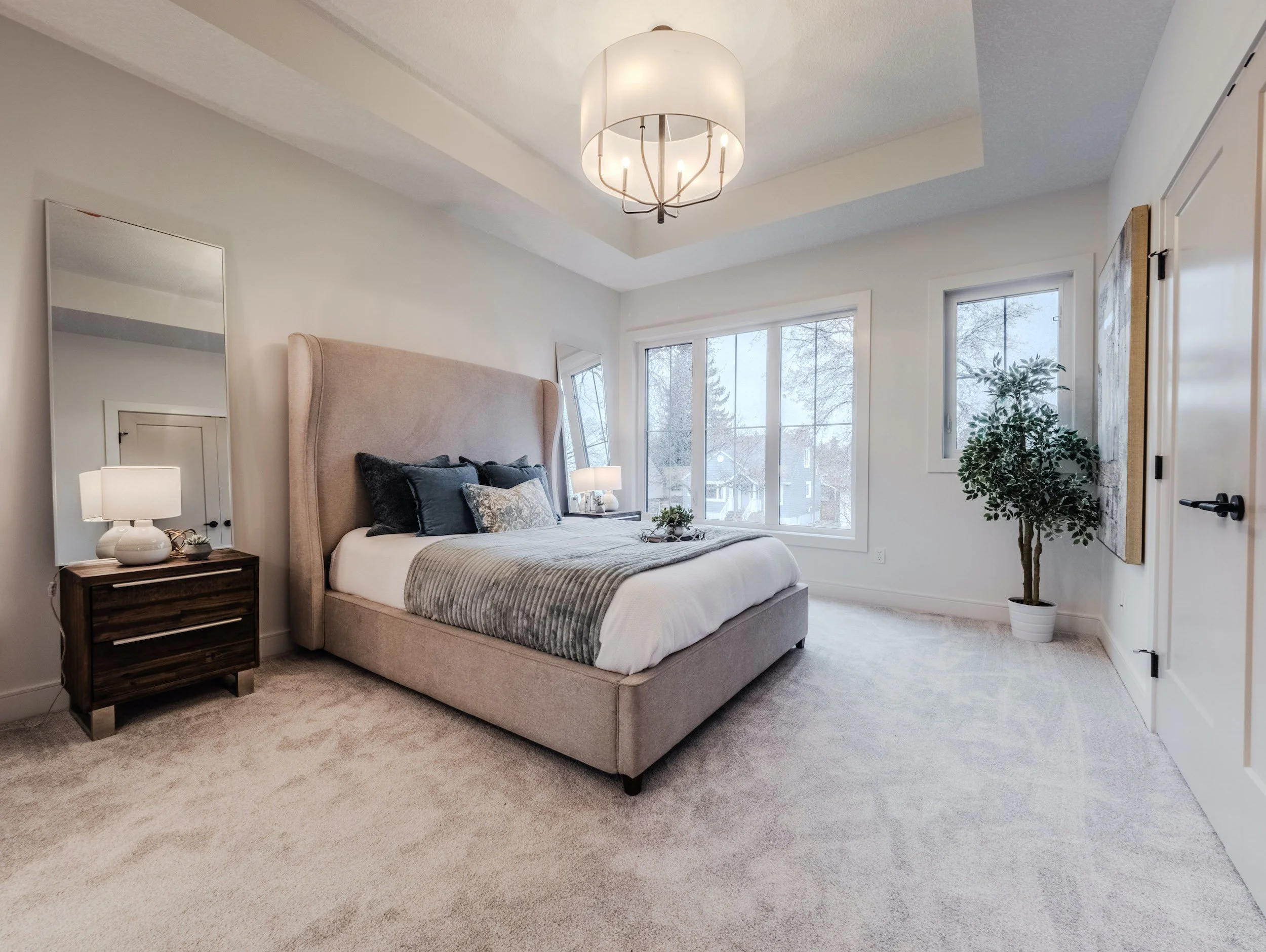 Serene primary bedroom with tray ceiling architecture, abundant natural light from large windows, and cozy, neutral-toned decor.