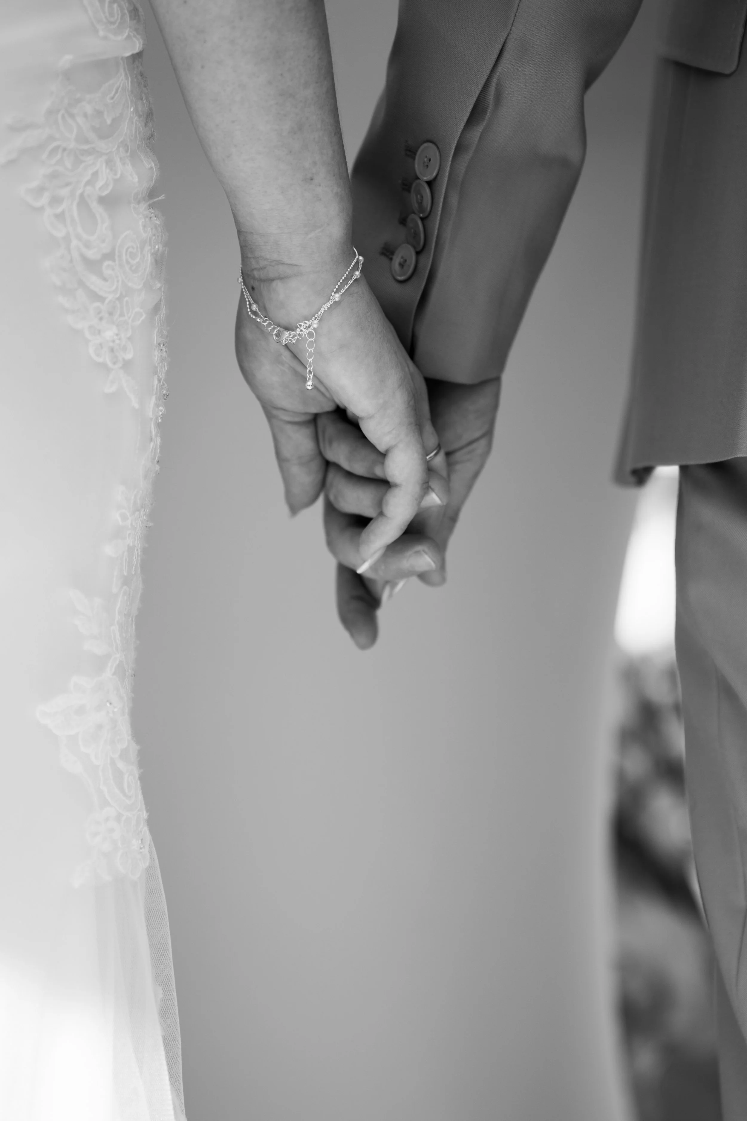 A close-up black-and-white photograph of a bride and groom holding hands during their wedding ceremony, with the bride wearing a lace wedding dress and a silver bracelet, and the groom dressed in a suit with buttons on the sleeve.