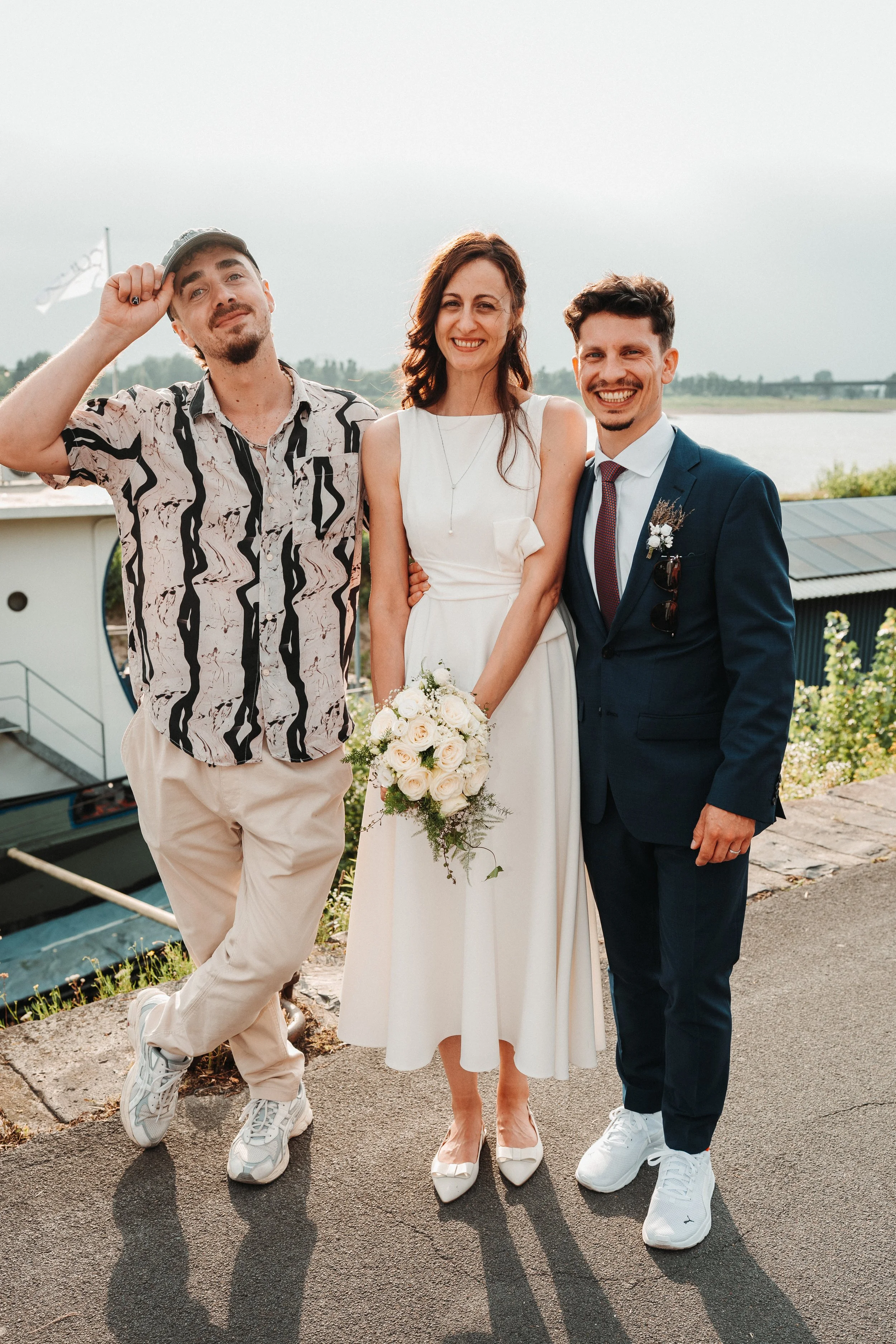 A bride and groom standing with a man outdoors near a body of water, smiling for a photo at a wedding.