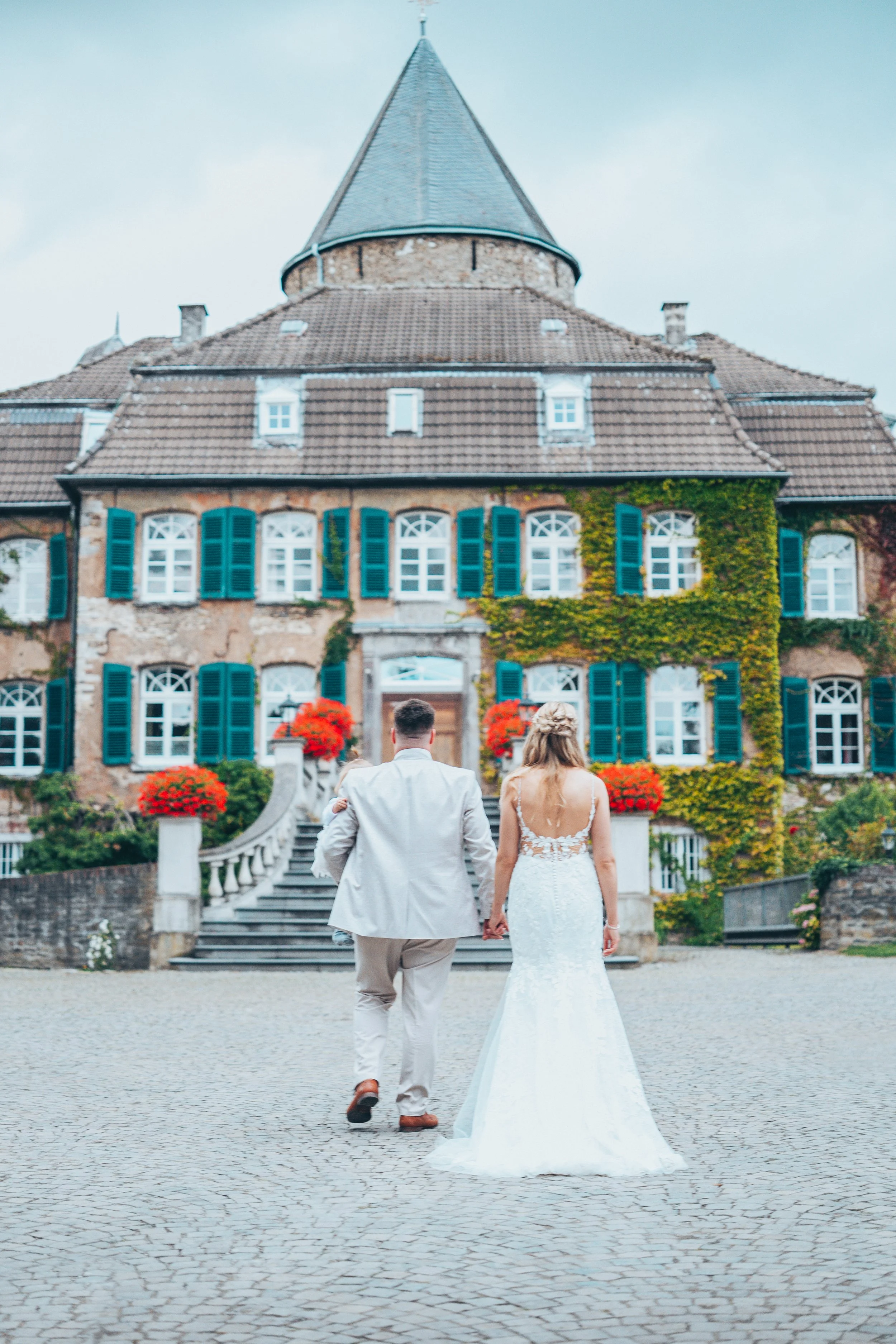 A bride and groom walking hand in hand towards a large, old mansion with ivy-covered walls, blue shutters, and a tall pointed roof, on a cobblestone driveway.