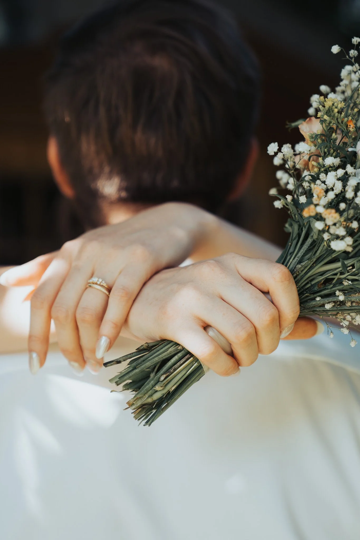 Nahaufnahme von einem Menschen, der eine kleine Blumenstrauß aus weißen und gelben Blumen hält, mit Händen, die eine goldene Ringschleife tragen.