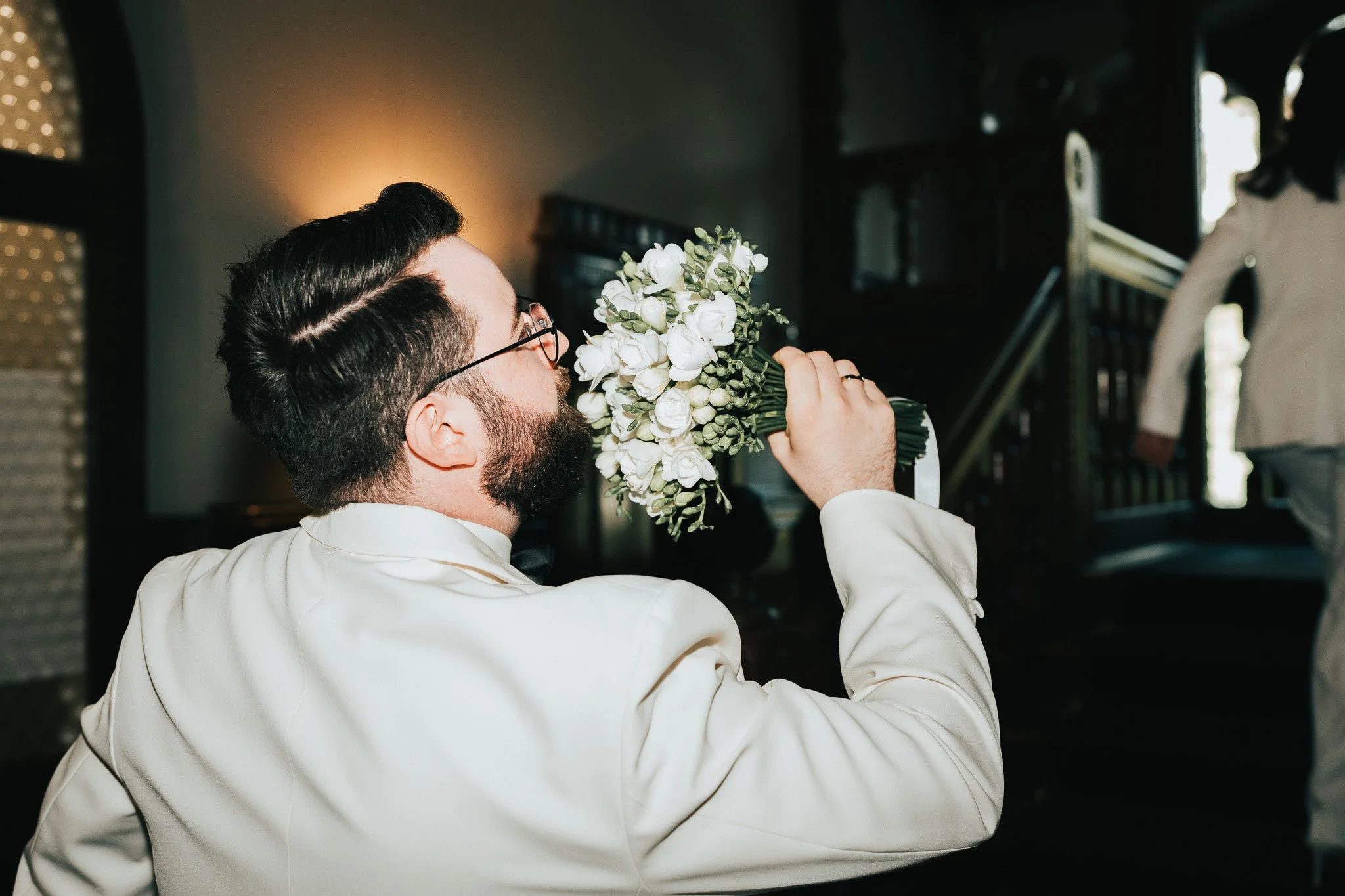 Ein Mann mit dunklen, gestylten Haaren, Bart und Brille hält ein weißes Blumenbouquet in der Hand.