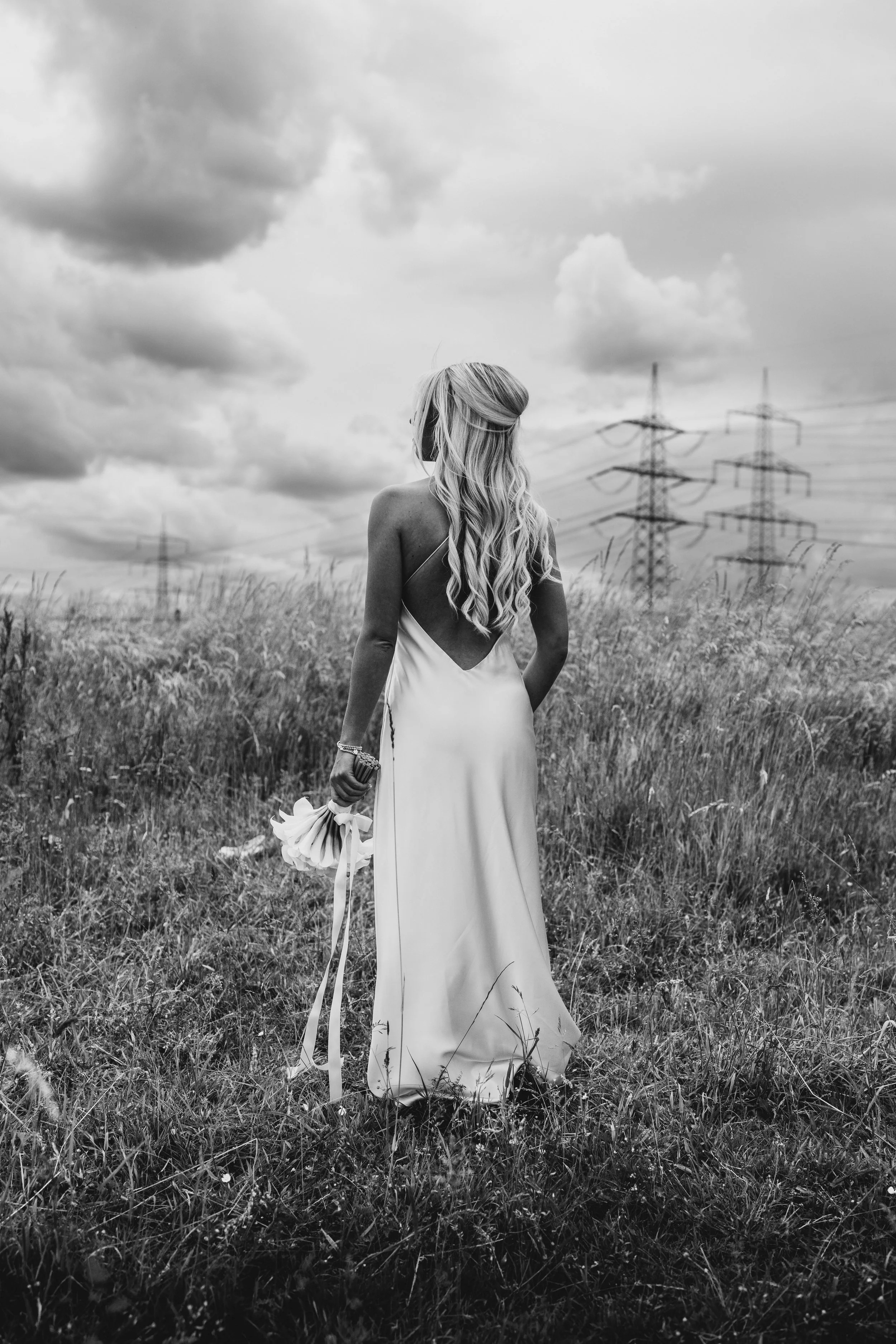 A woman in a long dress stands in a field holding a bouquet, with power lines and cloudy sky in the background, in black and white.