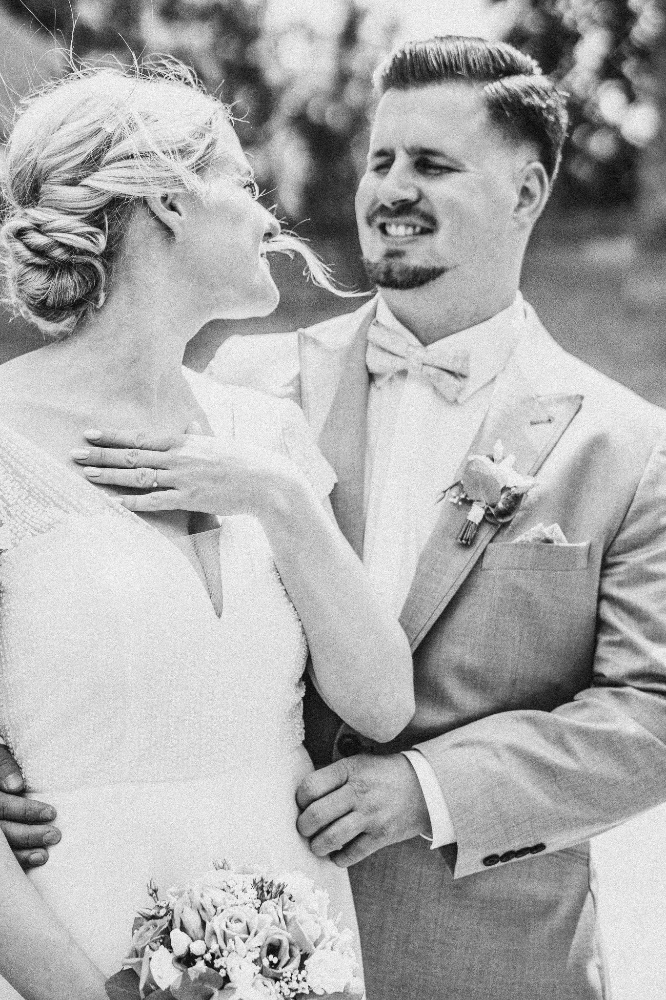 A black-and-white photograph of a bride and groom on their wedding day, with the groom looking at the bride and holding her waist. The bride is smiling, holding a bouquet, and has her hand on her chest. The groom is dressed in a suit with a bow tie a