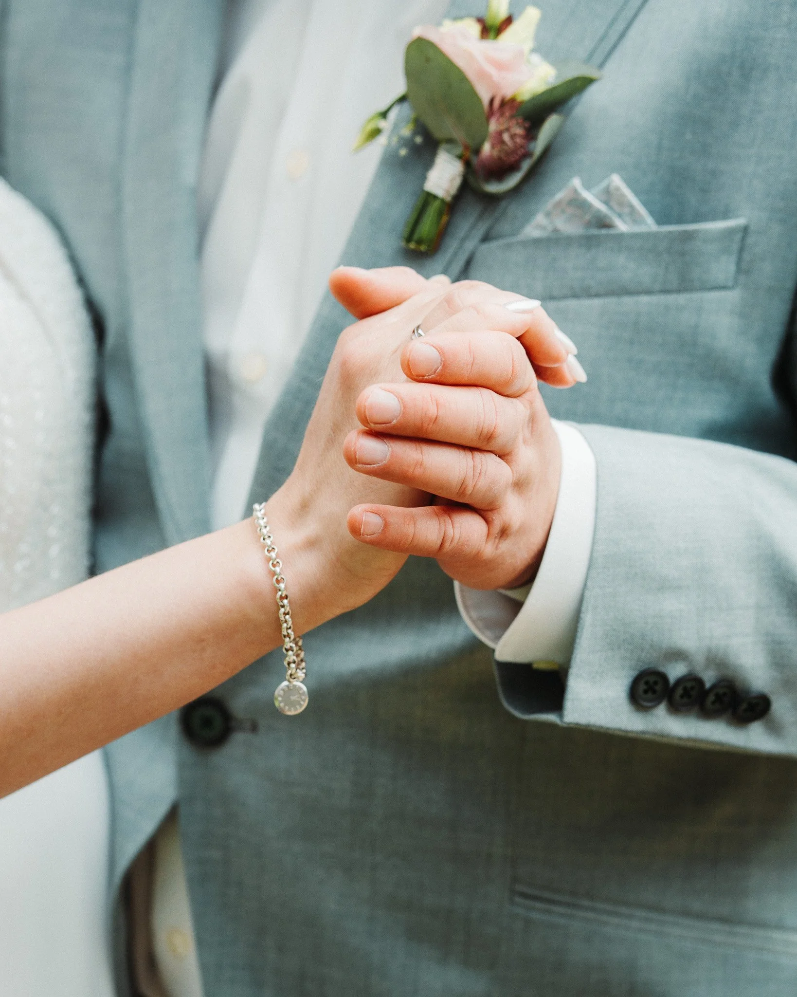 Close-up of a newlywed couple holding hands, with the bride wearing a bracelet and the groom in a light gray suit with a boutonniere.