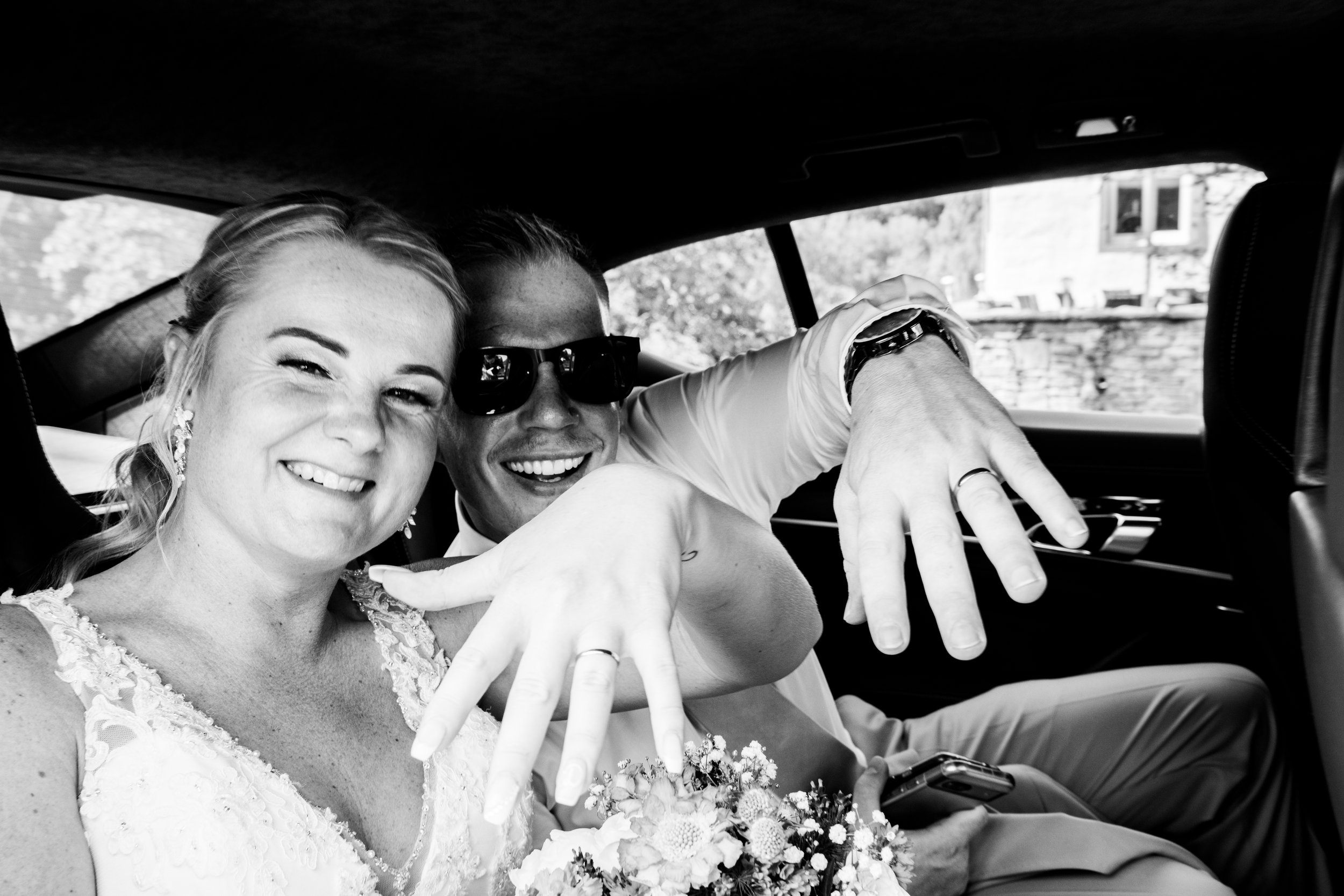 Black and white photo of a smiling couple in wedding attire sitting in a car, celebrating their wedding day.