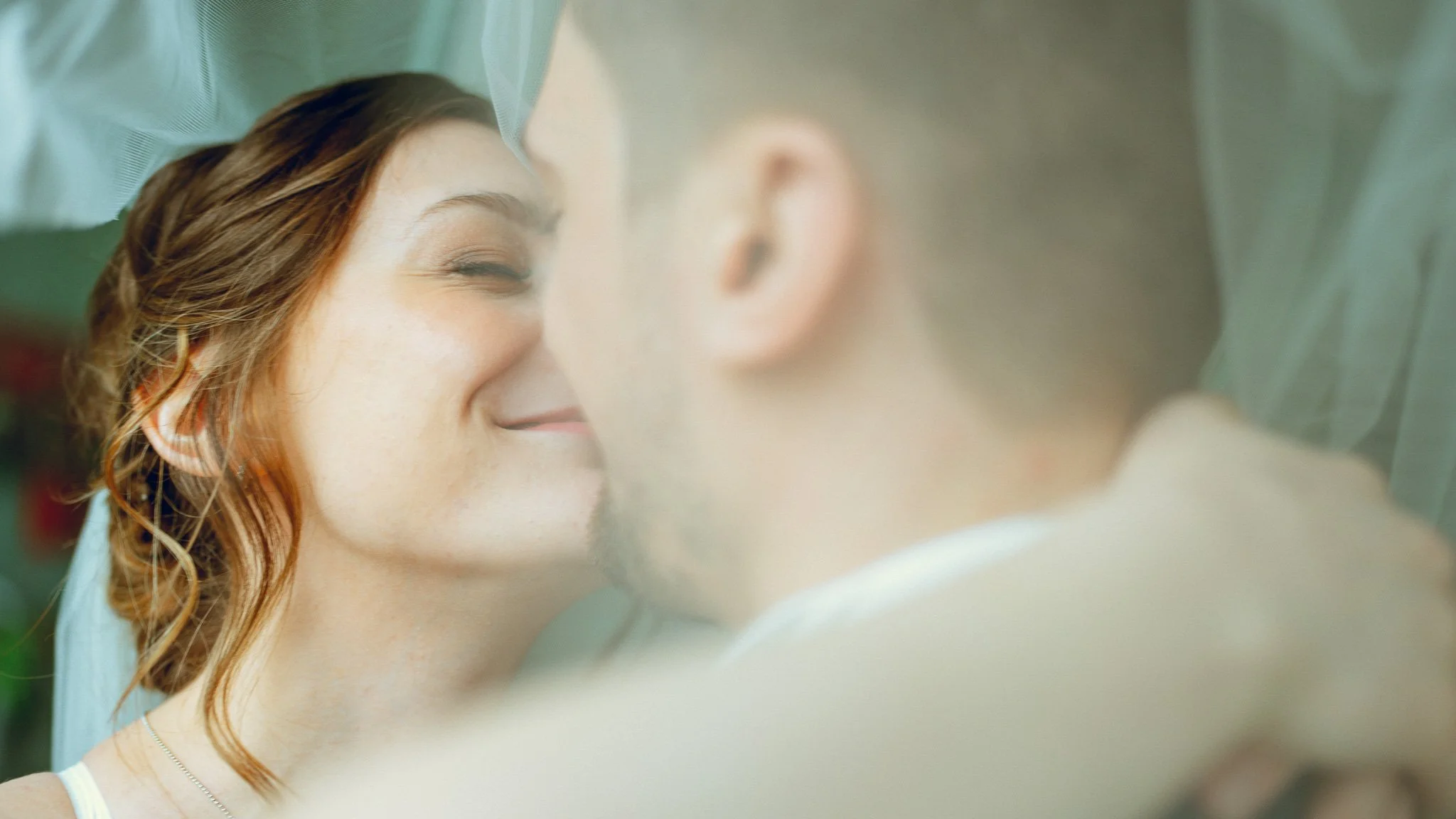 A woman with auburn hair smiling with her eyes closed, close to a man with short light hair and beard, both under a translucent veil, with an intimate and joyful expression.