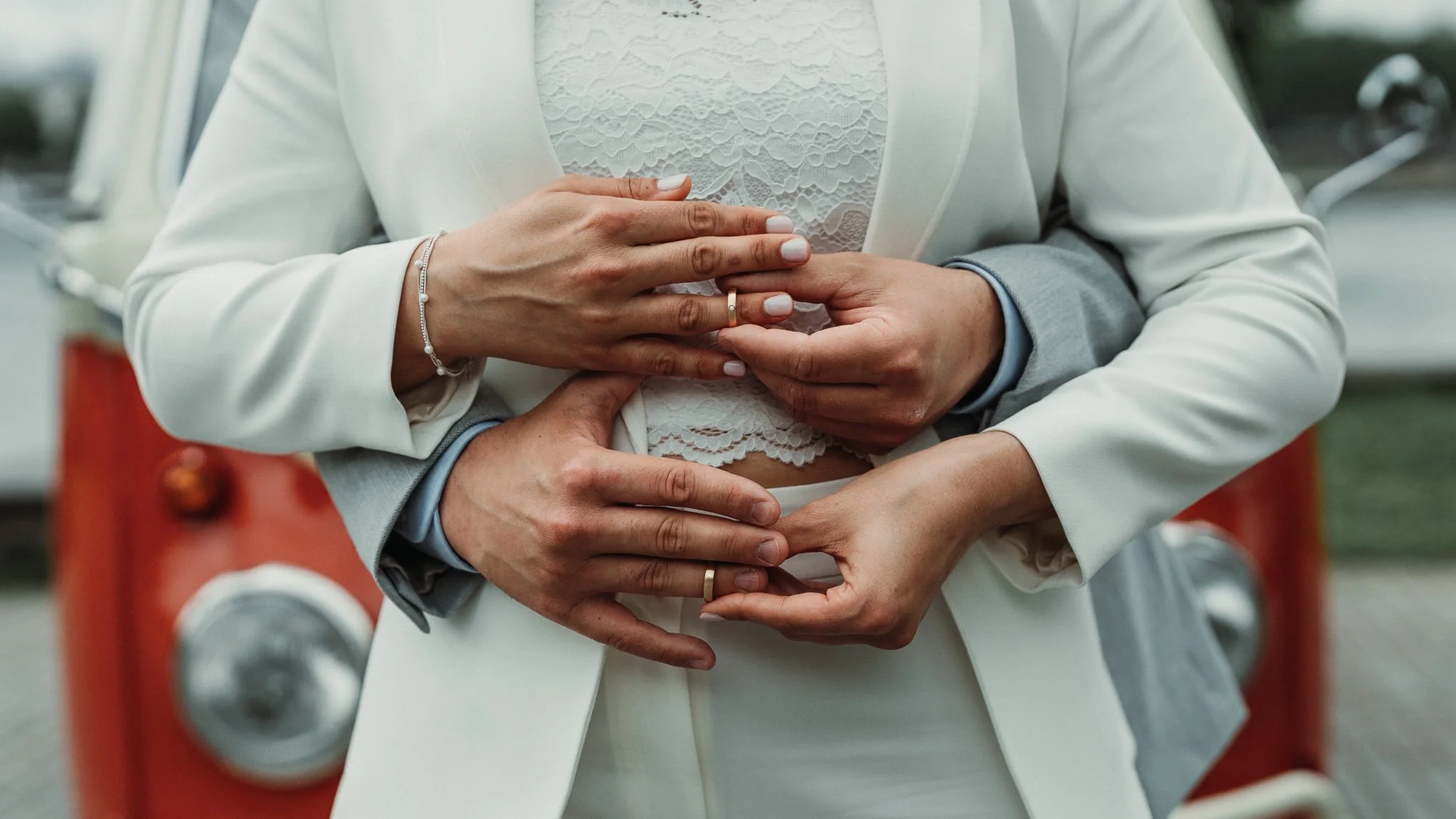 Close-up of a bride and groom holding hands, displaying wedding rings, with the bride on top and groom below, with a vintage car in the background.