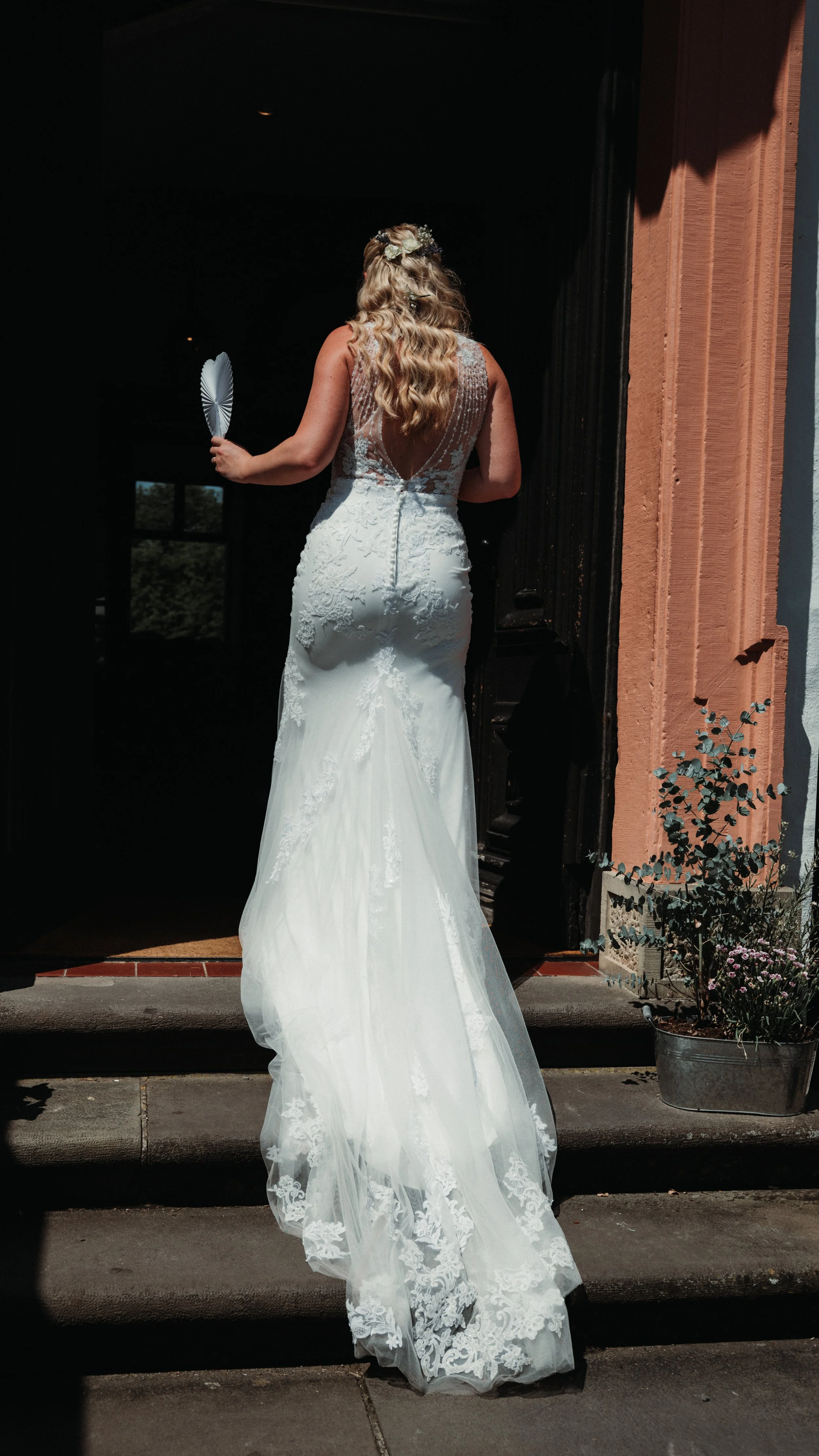 Back view of a bride in a white lace wedding gown holding a fan, standing on a porch with steps leading up to a doorway, with sunlight casting shadows and potted plants nearby.