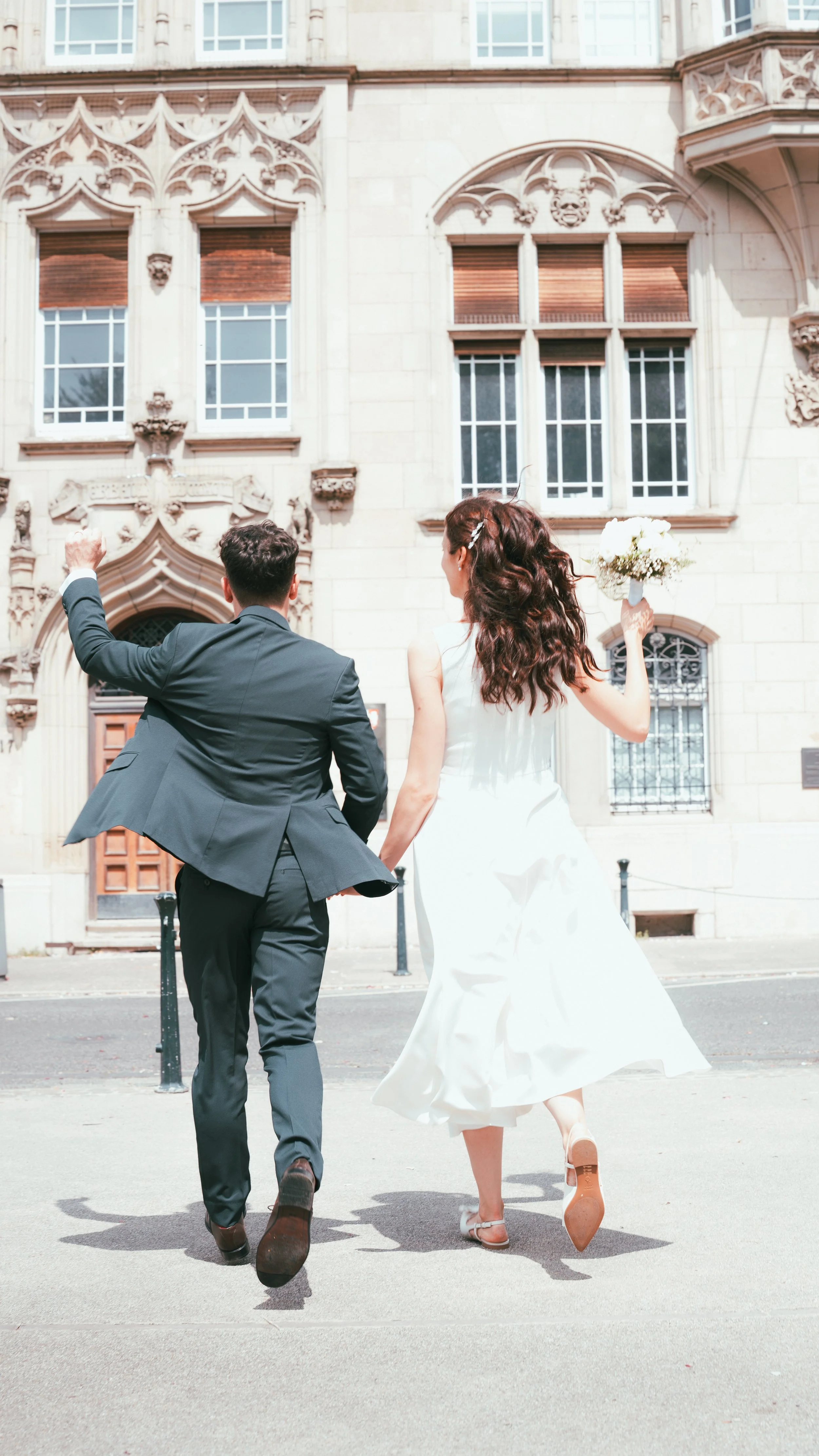 Couple in wedding attire walking hand-in-hand on a city street, with the groom in a suit and the bride in a white dress holding a bouquet, approaching a historic building.