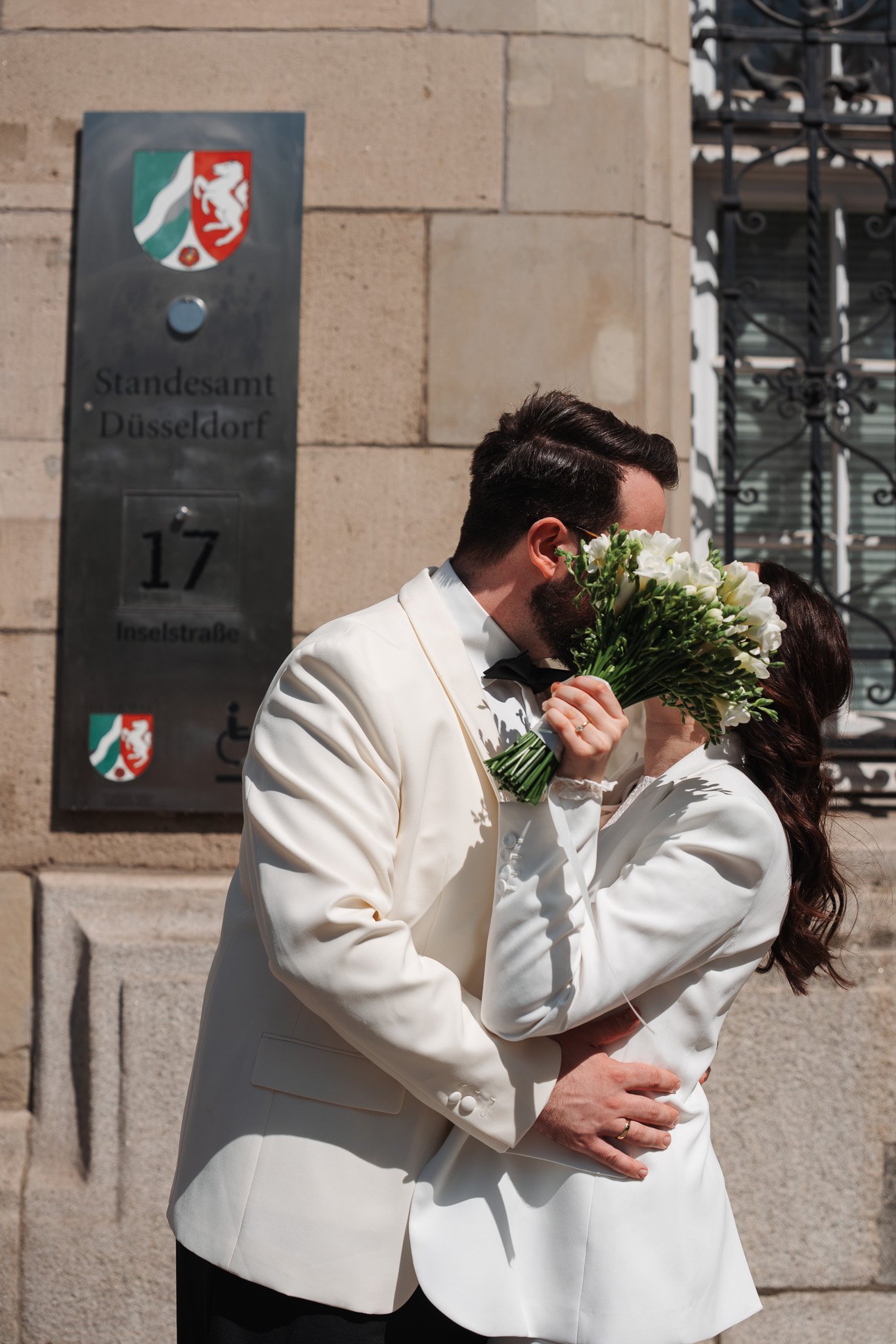 Ein Mann und eine Frau, beide in weißen Anzügen, umarmen sich vor einem Gebäude mit einem Schild, das auf das Standesamt Düsseldorf hinweist. Der Mann hält einen Blumenstrauß.