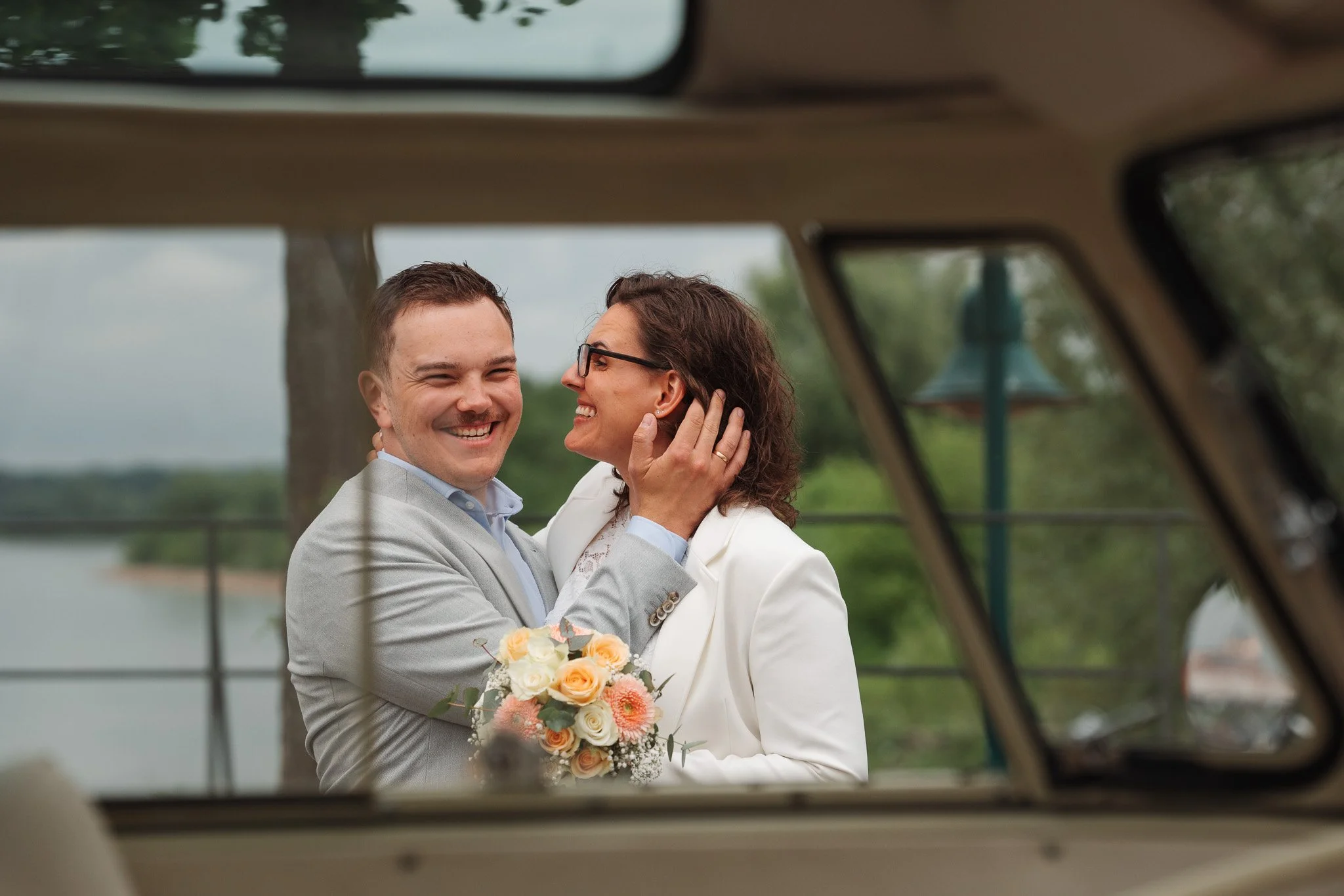 A smiling couple, dressed formally, embracing outdoors with a bouquet in hand, viewed through a window or opening with a blurred background of trees and water.