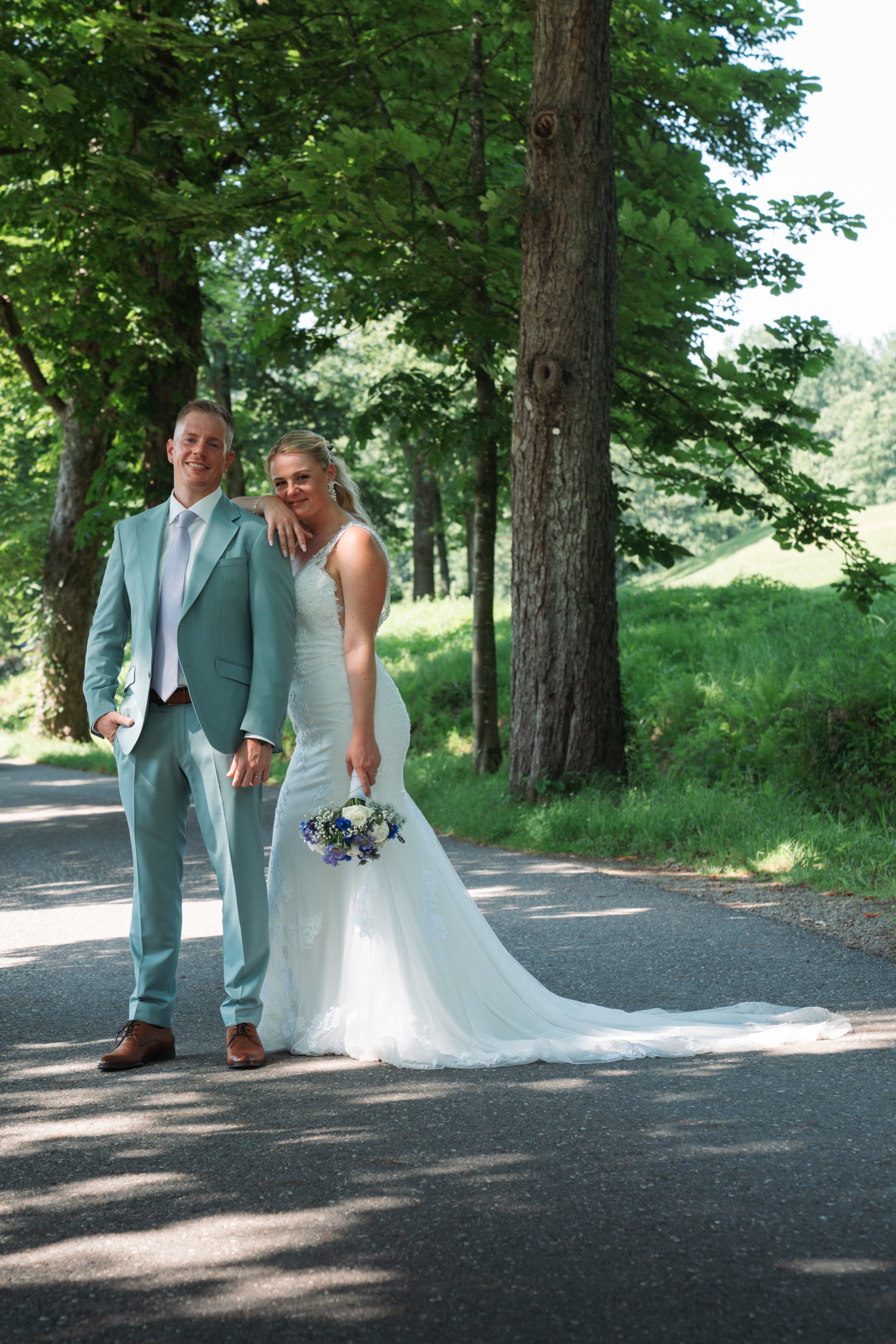A bride and groom standing on a shaded road in a wooded park during daytime, smiling for the camera. The bride is in a white wedding dress holding a bouquet, and the groom is in a light blue suit with a white shirt and tie.