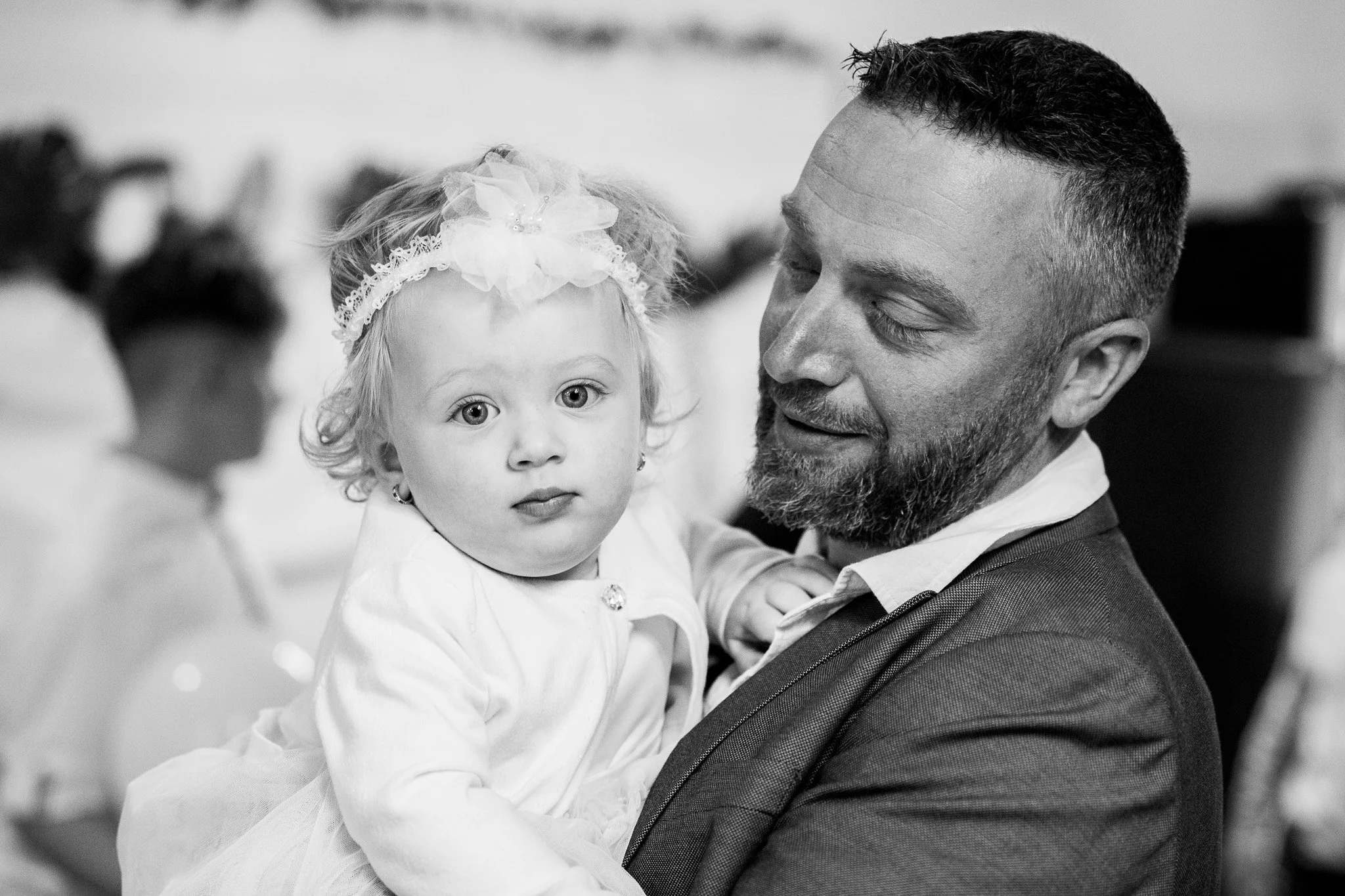 A man with a beard holding a young girl with curly hair, a headband, and a dress, at a likely indoor social gathering, captured in black and white.