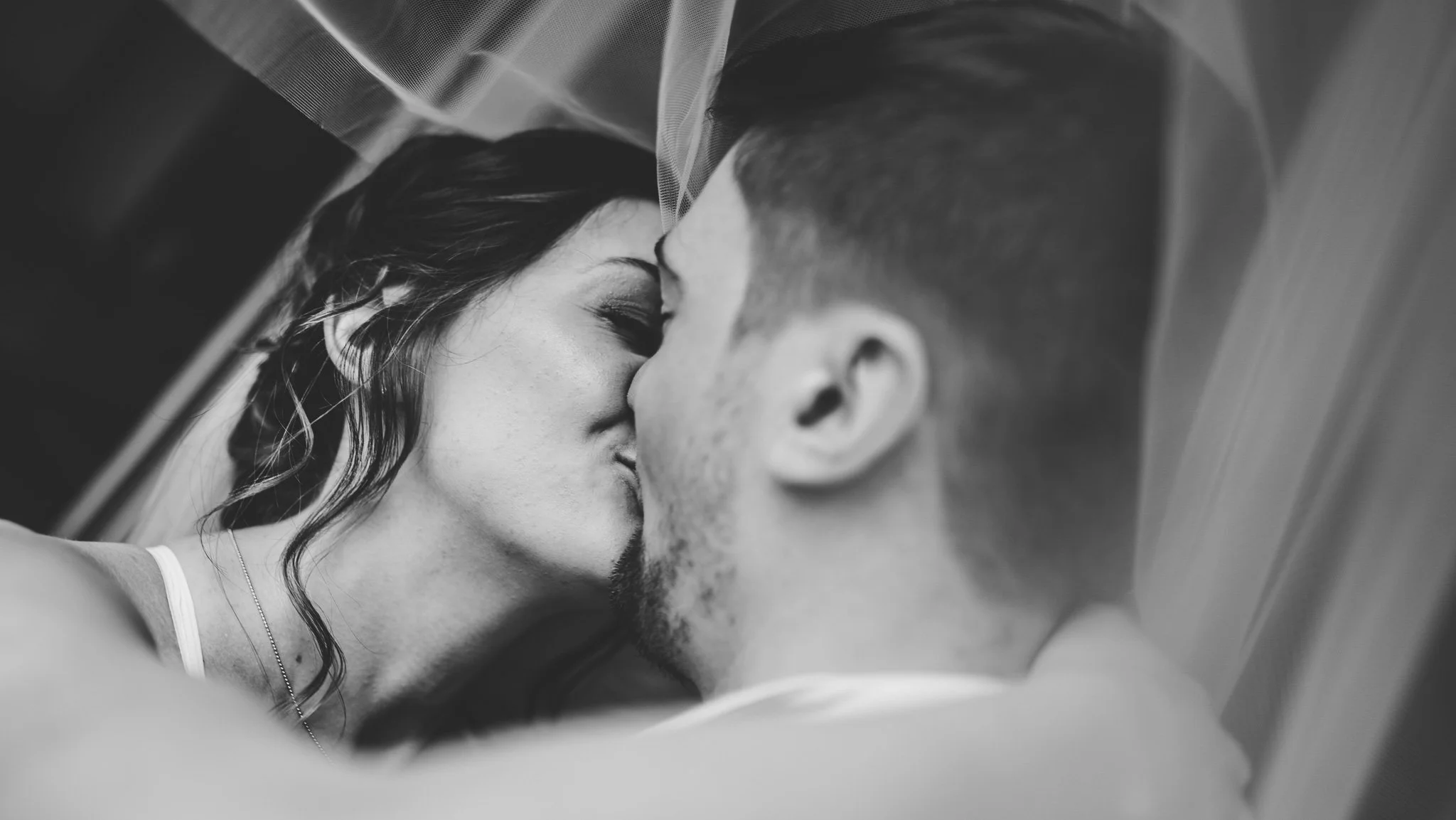 A black and white photograph of a man and woman lying close together, about to kiss, with their faces touching and eyes closed.