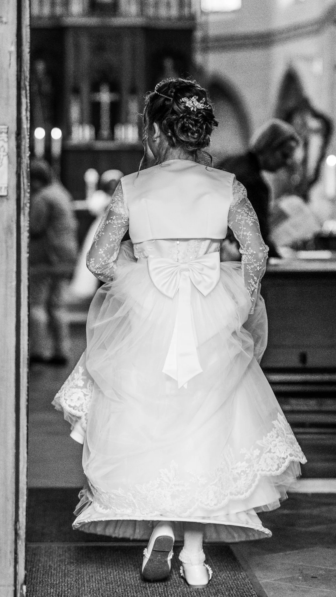 A young girl dressed in a formal gown with lace sleeves, a large bow on the back, and wearing dress shoes, walking inside a church or chapel.