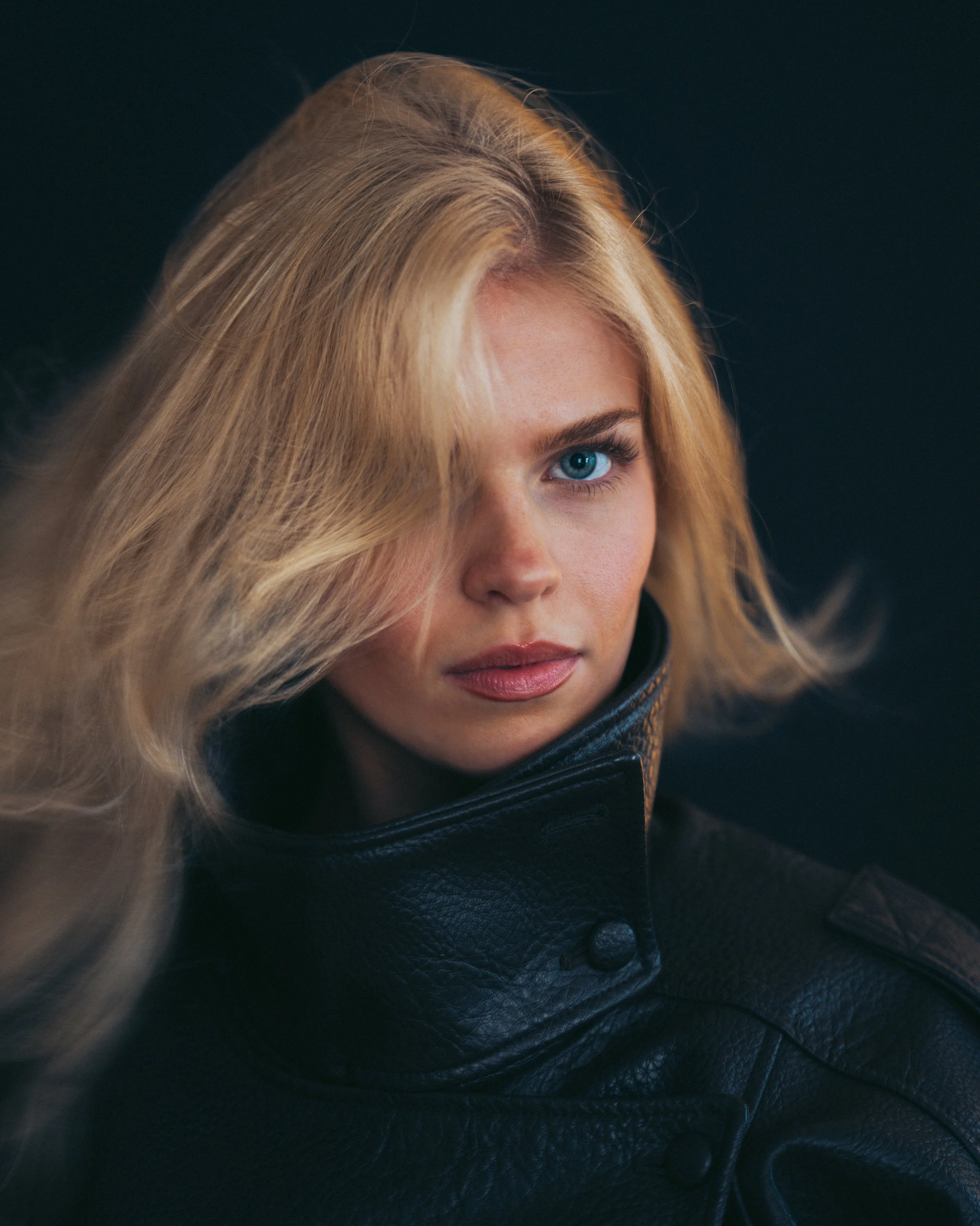 Close-up of a woman with blonde hair and blue eyes, wearing a dark leather jacket, against a dark background.