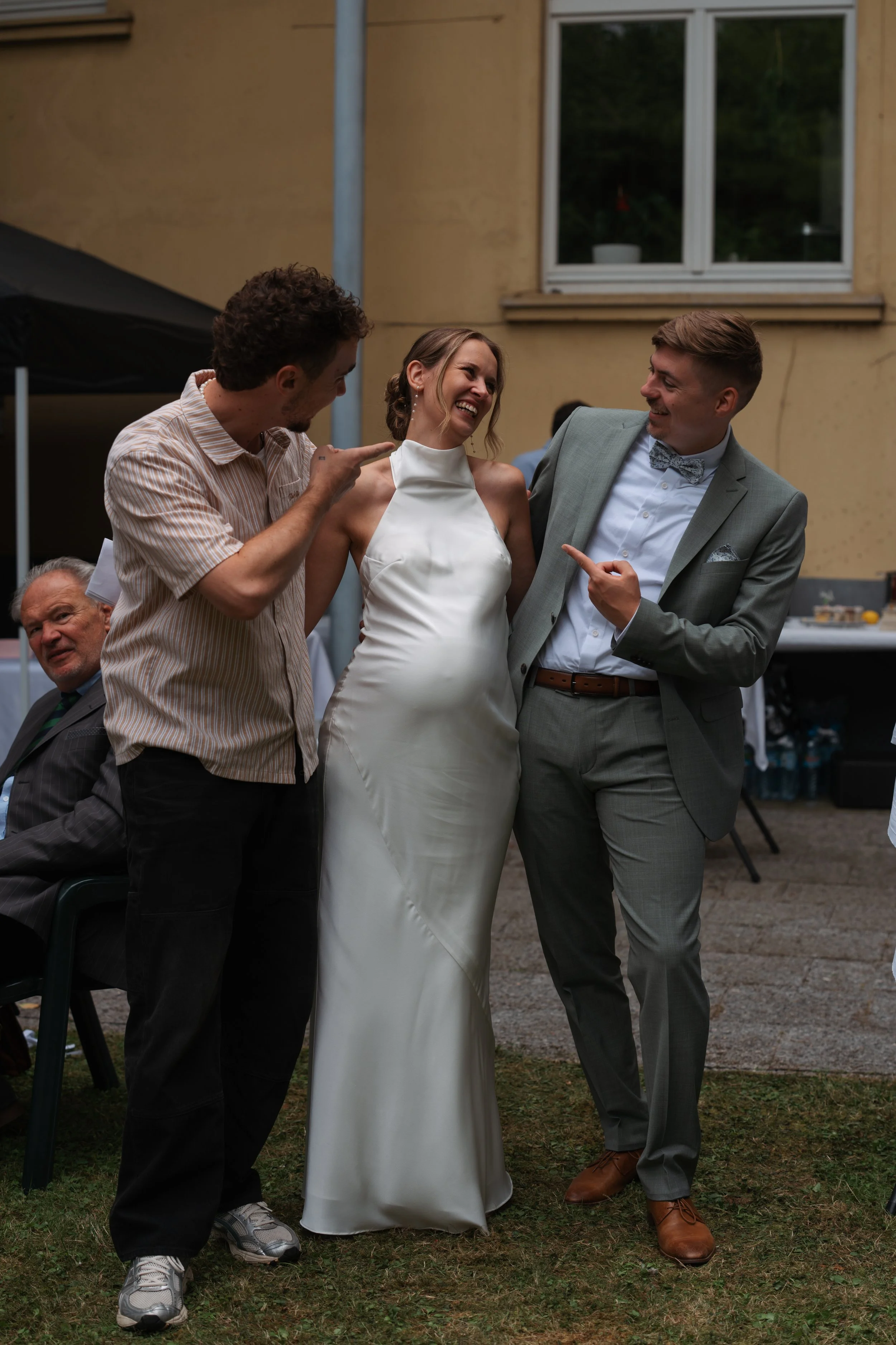 Three people at a gathering are enjoying a conversation, with a woman in a white dress smiling and flanked by two men in suits, one pointing and the other smiling, outdoors with a yellow building and a window in the background.