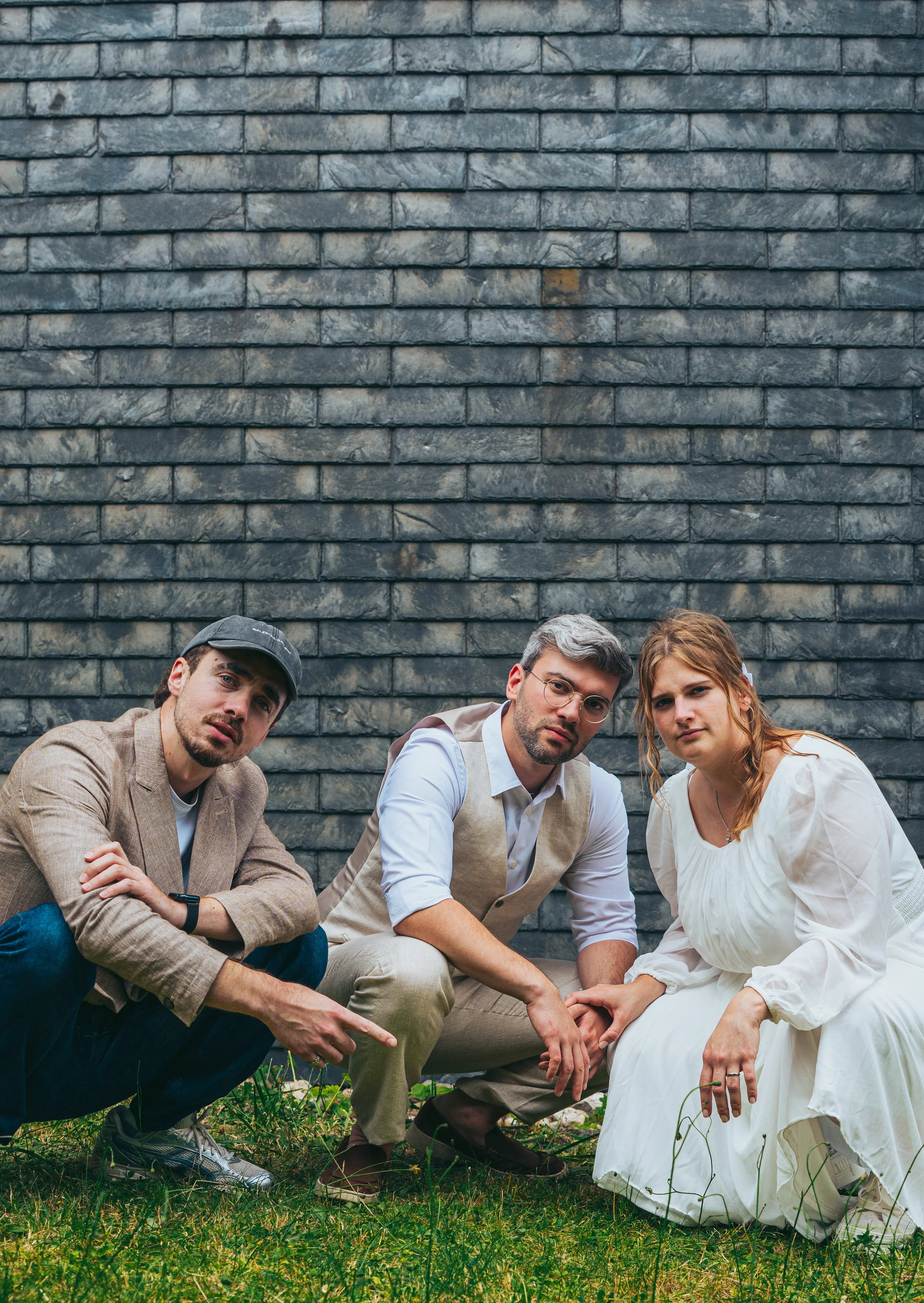 Four people crouching and sitting in front of a brick wall, surrounded by green grass and small plants.