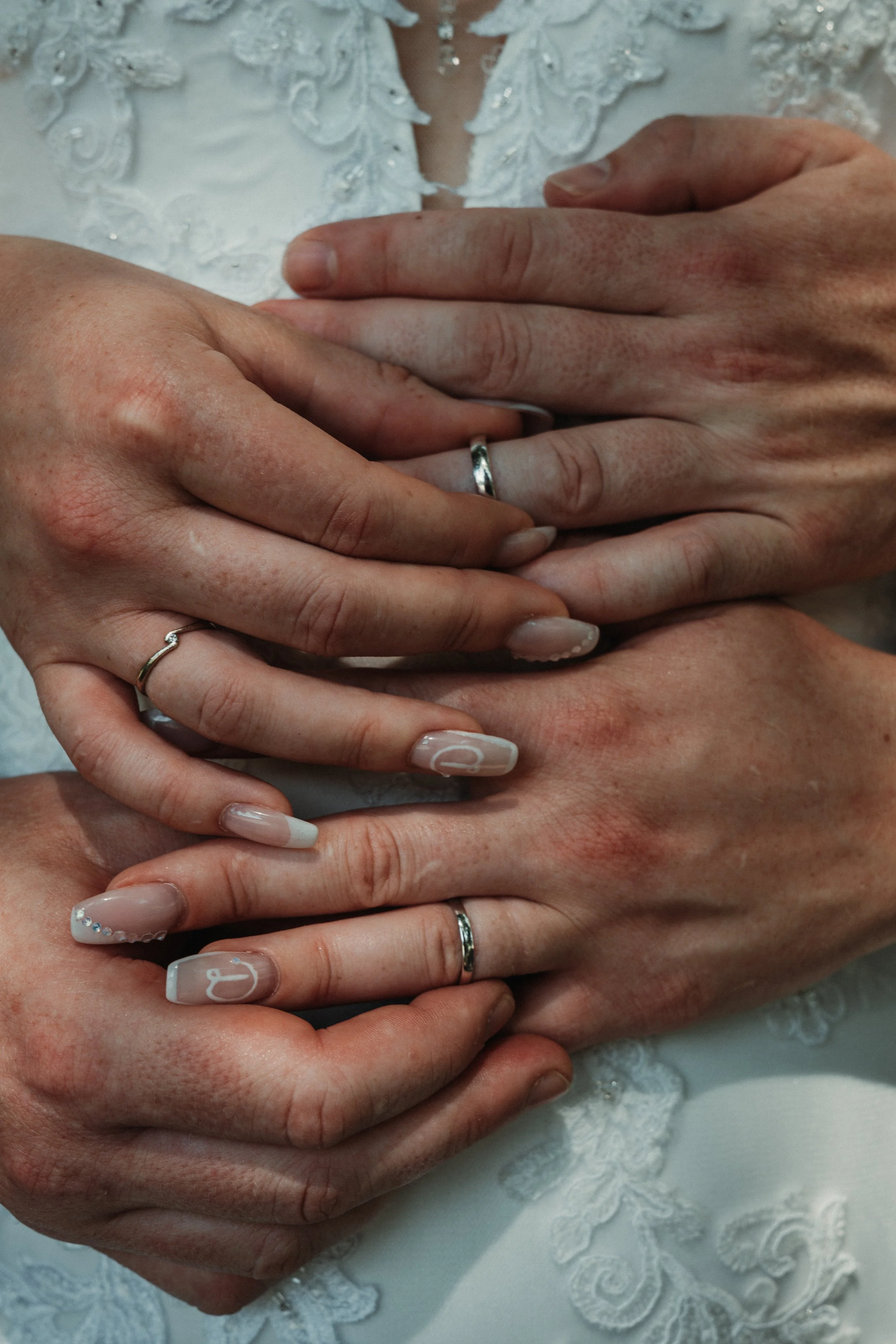 Multiple hands with wedding rings placed on each other's palms, in front of a white lace dress, symbolizing marriage or commitment.