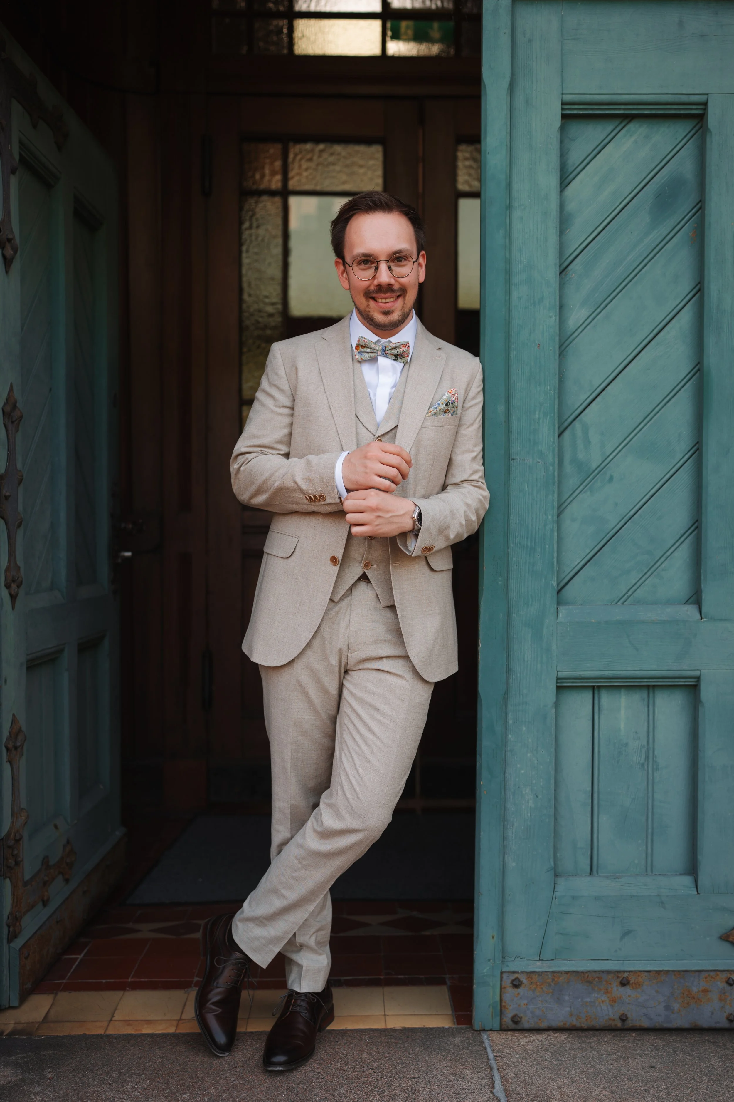 A man wearing a beige suit, white shirt, floral bow tie, and glasses, standing partially inside a teal barn door, smiling at the camera.