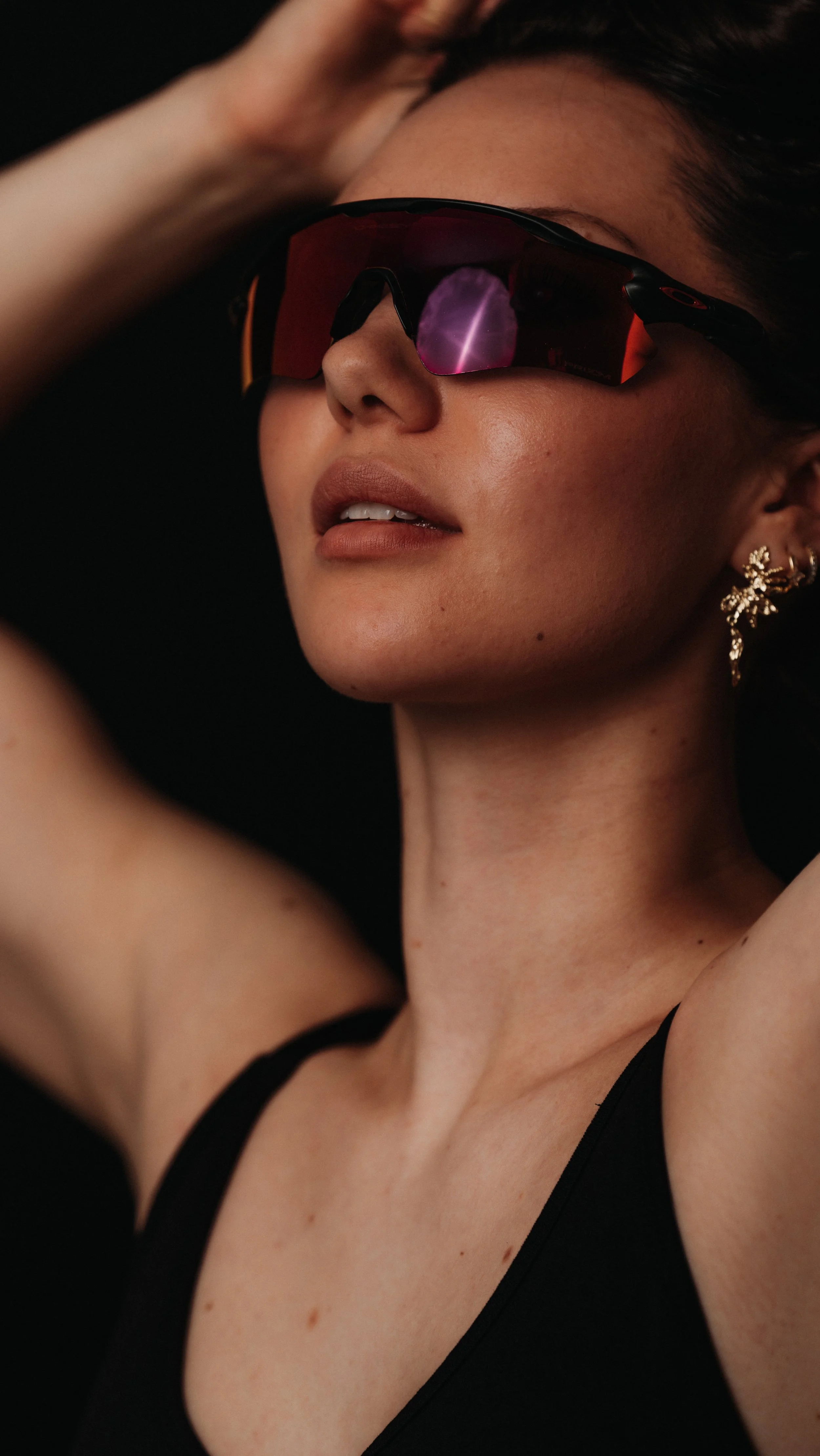 A woman with short dark hair wearing sunglasses with red reflective lenses, gold earrings, and a black top, against a black background.
