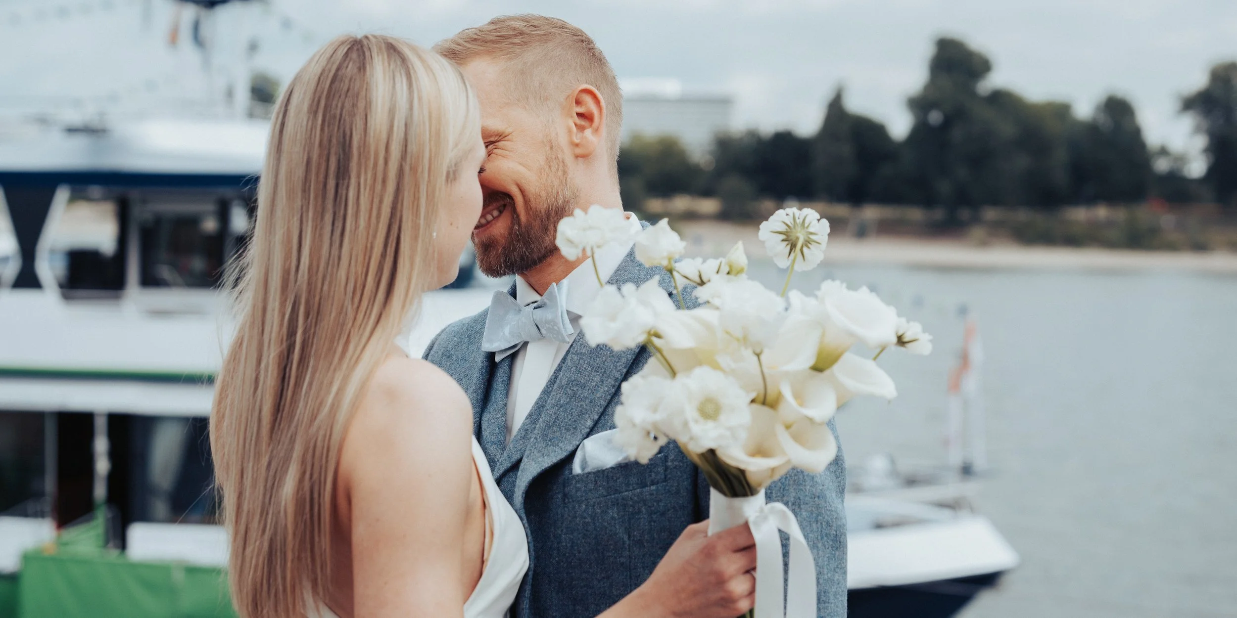 A couple dressed in wedding attire, smiling and leaning their foreheads together by a body of water, with a woman holding a bouquet of white flowers.