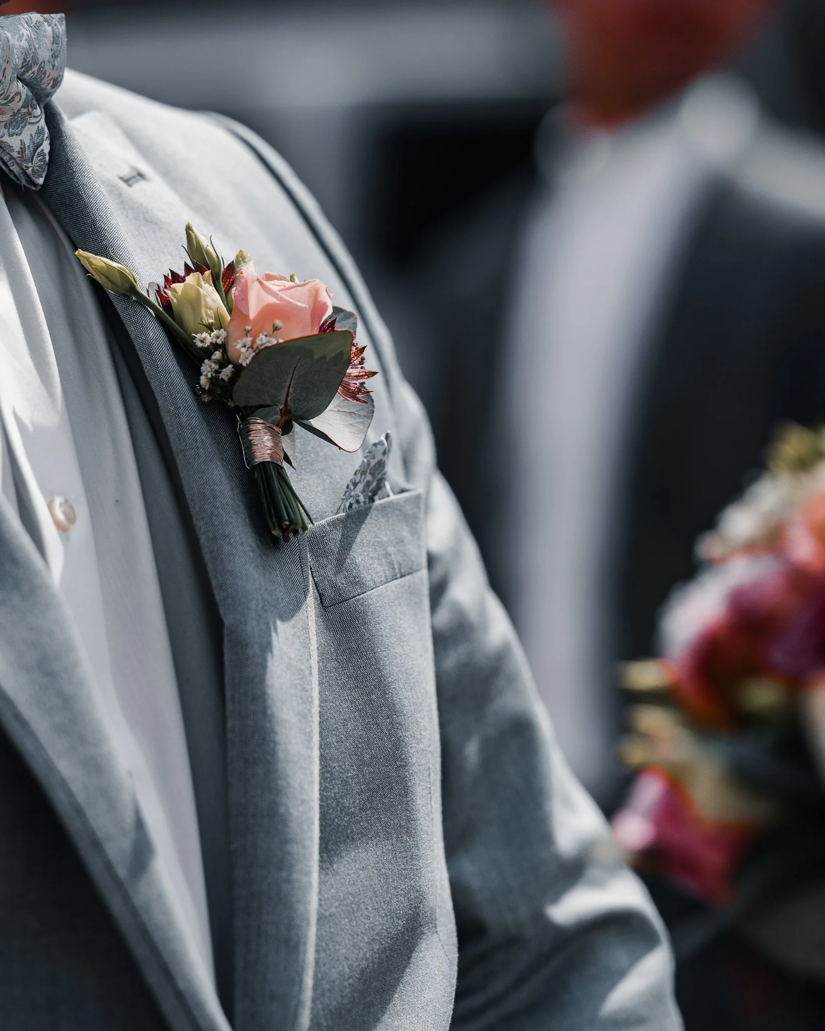 A close-up of a groom in a gray suit with a boutonniere consisting of a pink rose, white flowers, dark leaves, and greenery, at a wedding.