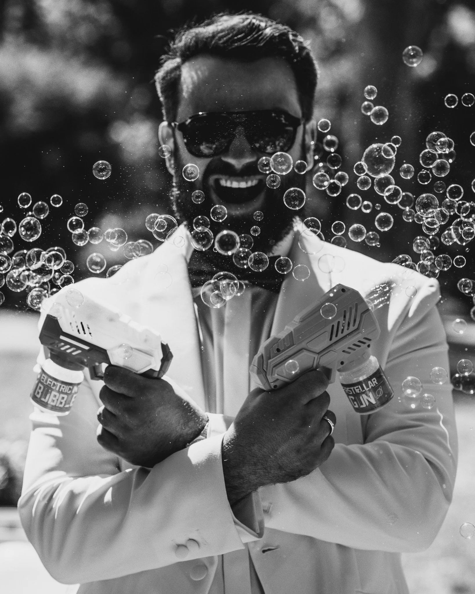 Schwarze Sonnenbrille, lachender Mann mit Bart, feuchte Haut, Sektflasche in der Hand, in einer Sanduhr-ähnlichen Pose, im weißen Anzug, im Freien, mit Seifenblasen, in Schwarz-Weiß