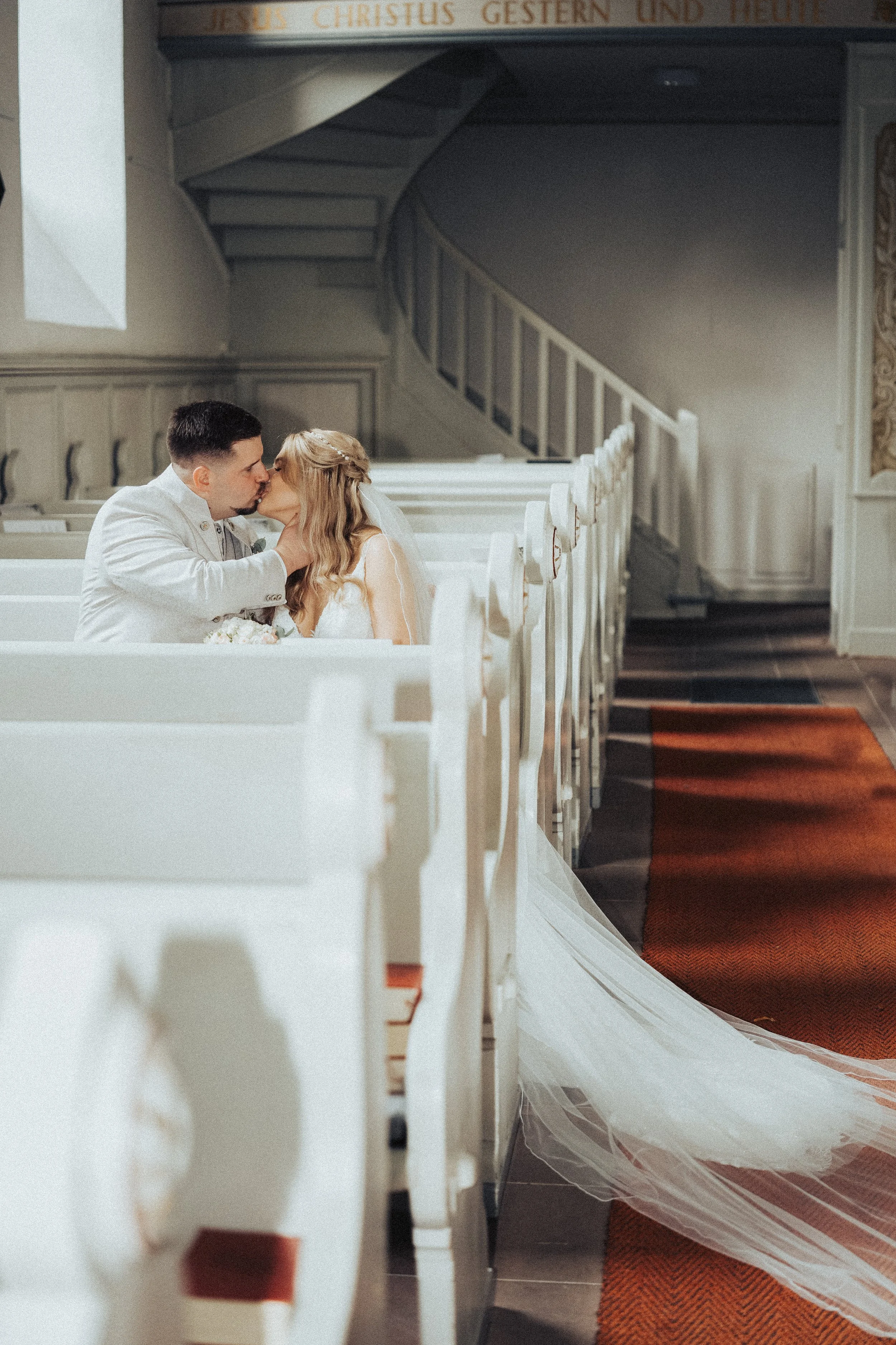 A bride and groom sharing a kiss inside a church pew during their wedding ceremony.