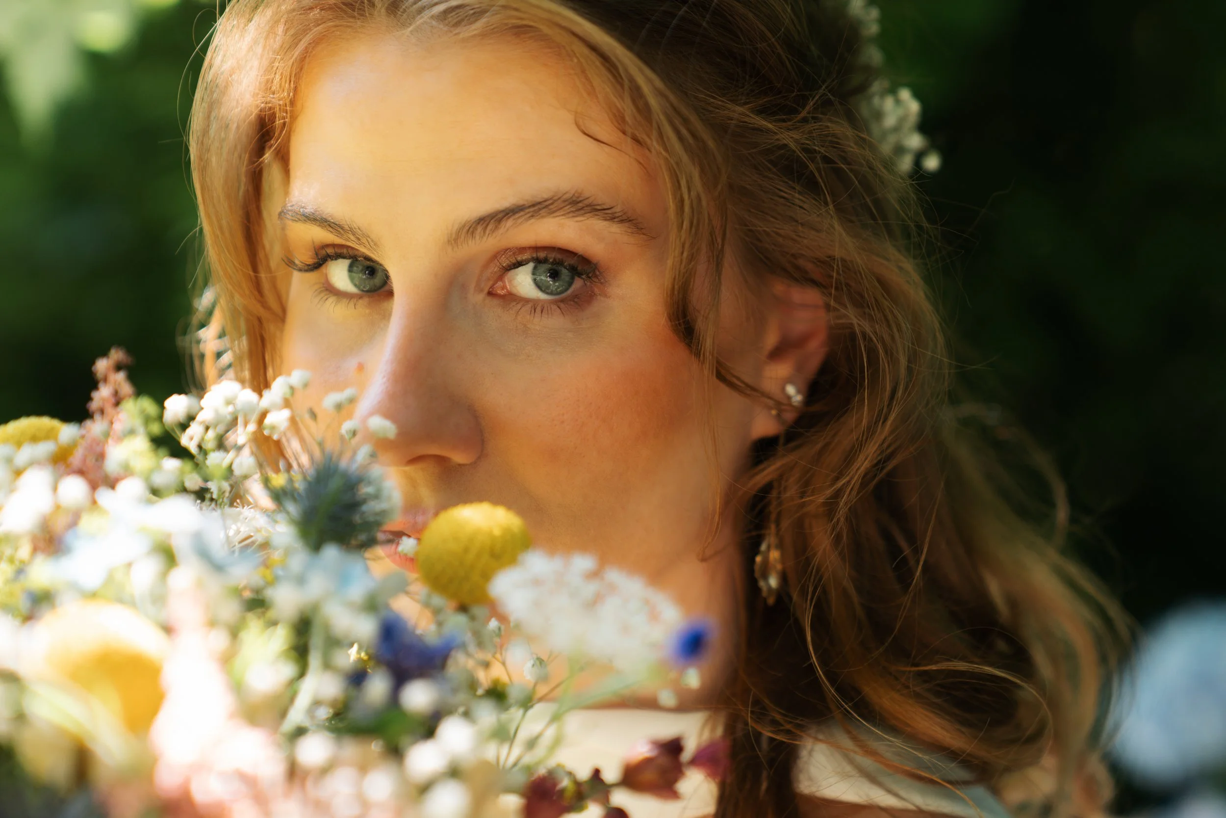 Close-up of a woman with green eyes and brown hair, surrounded by colorful flowers outdoors.
