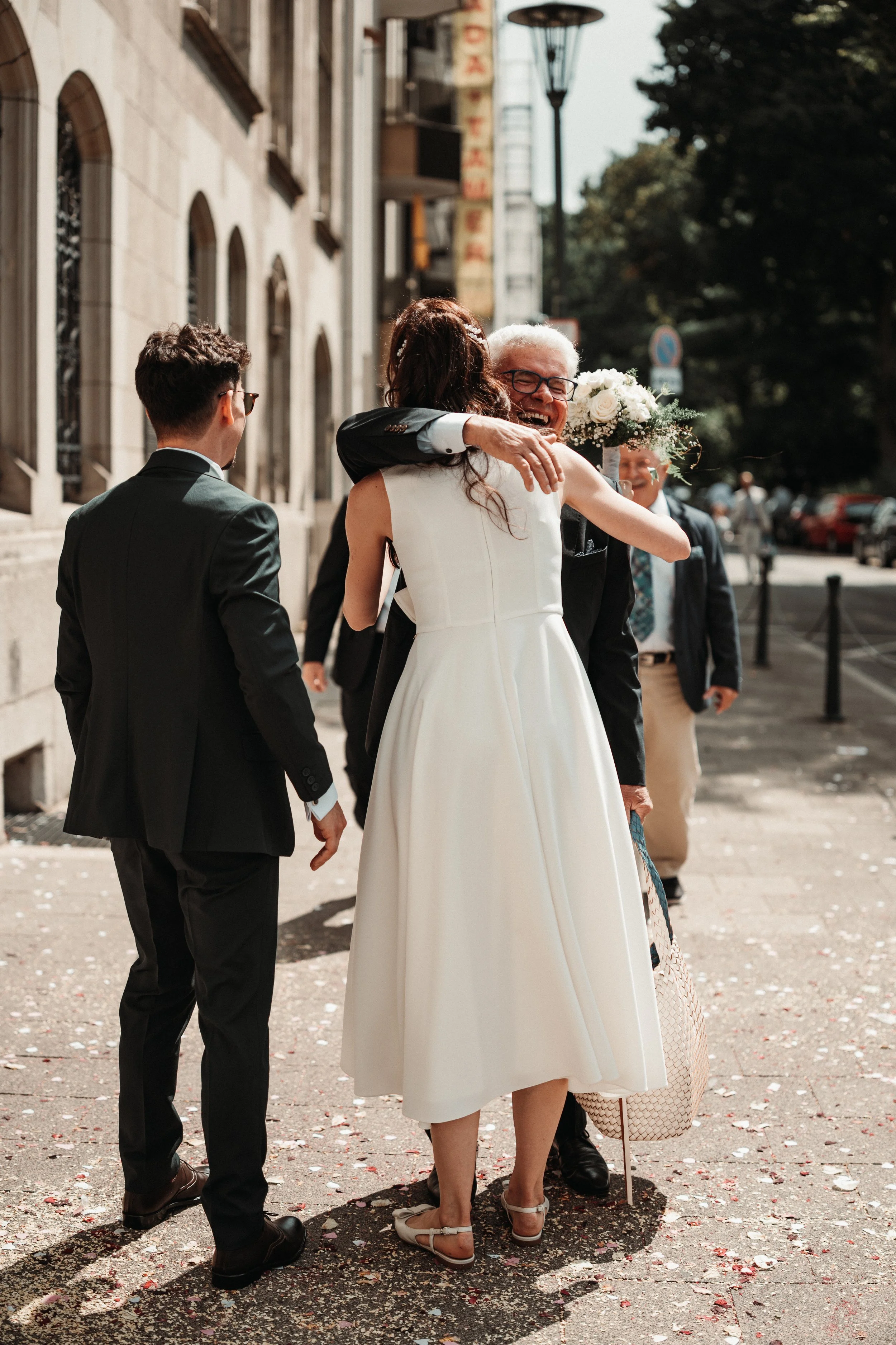 A joyful woman in a white dress hugging an older man with glasses, holding a bouquet of white flowers, with other people in formal attire around them, on a city sidewalk.