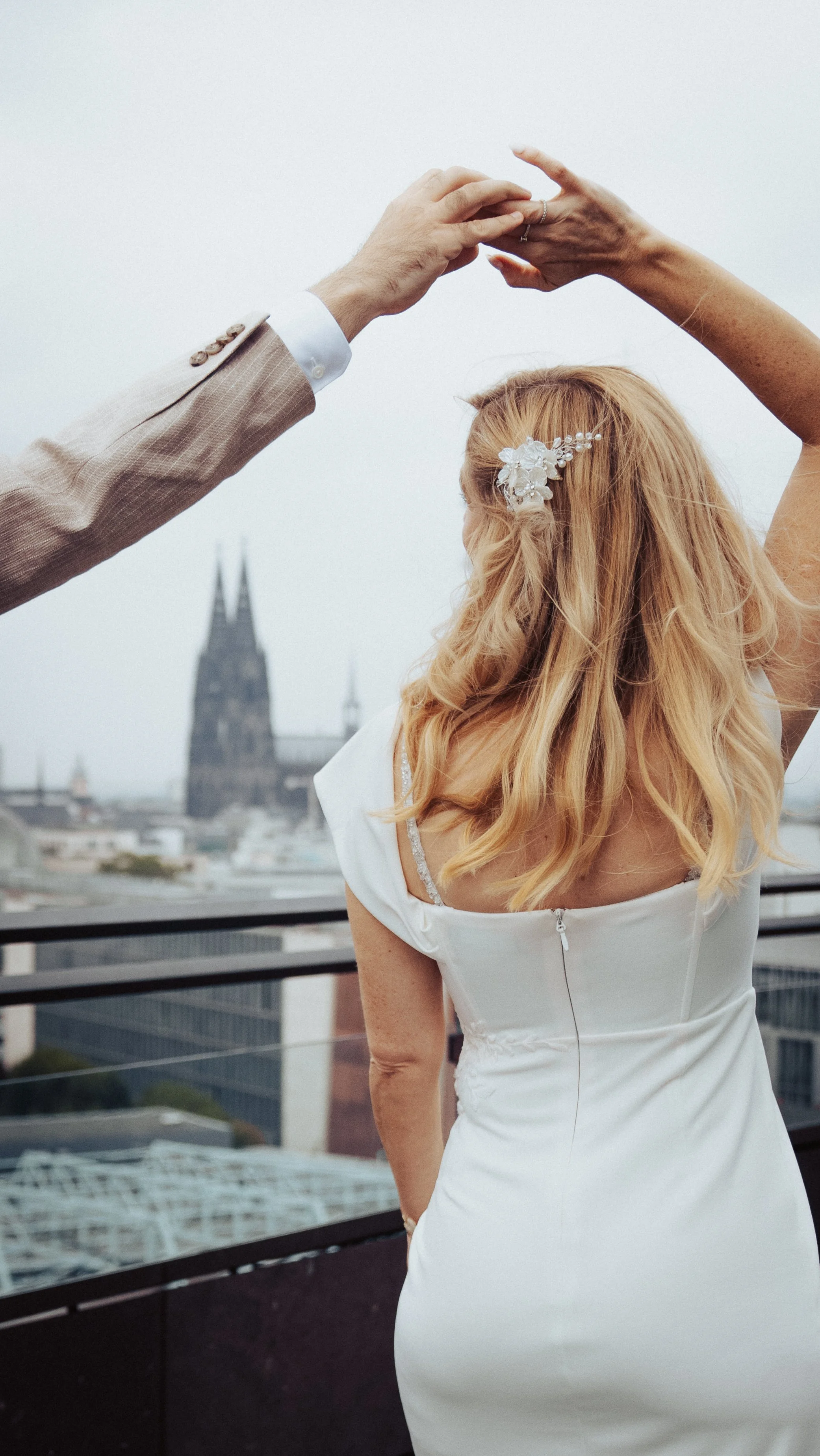 A bride and groom holding hands overhead on a rooftop with a city skyline and church spires in the background.