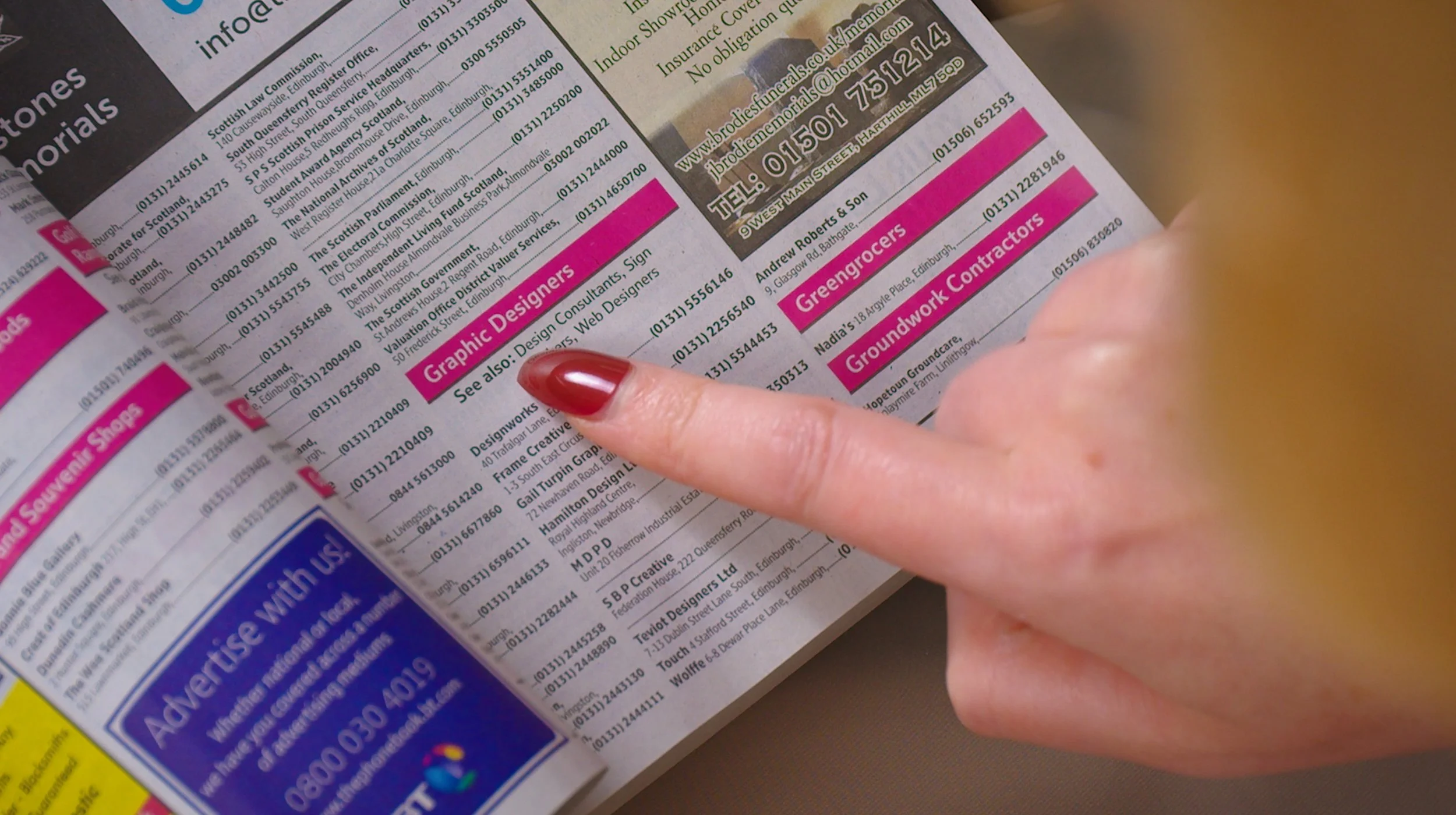 A person's hand with red painted nails points to a section of a newspaper or magazine, specifically towards an advertisement or listing titled 'Graphic Designers'.