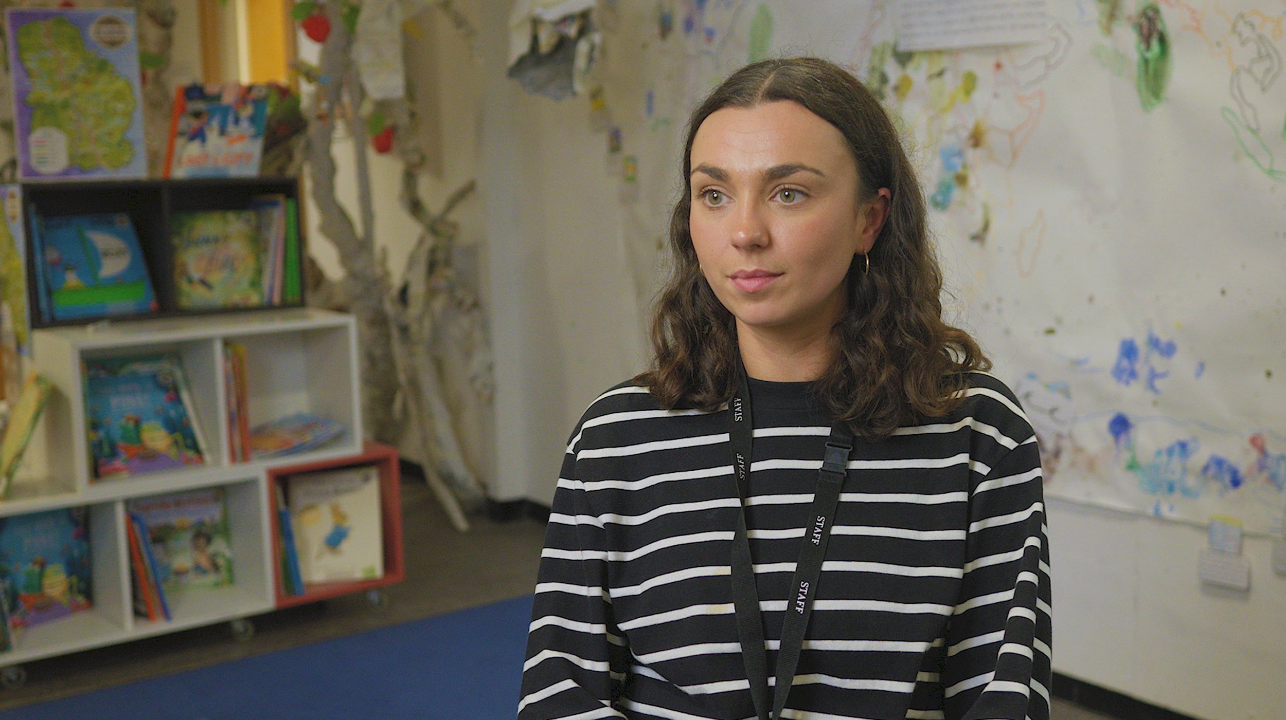 A young woman with shoulder-length wavy brown hair, wearing a black and white striped shirt and a black lanyard, stands in a colorful classroom with children's books and children's art on the walls.