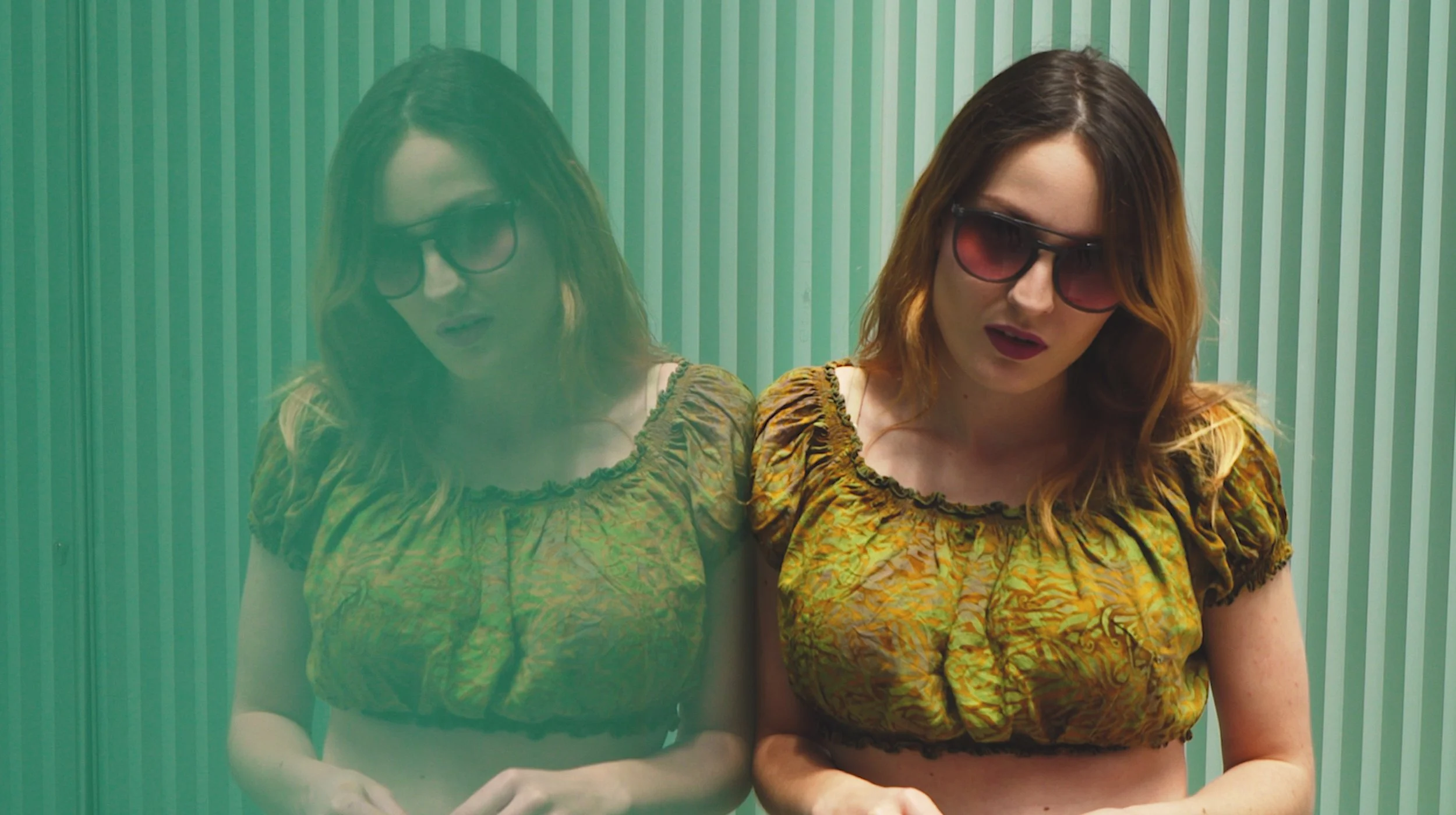A woman with red hair wearing sunglasses and a patterned yellow-orange top stands against a striped green background, with a semi-transparent reflection of herself on the left.