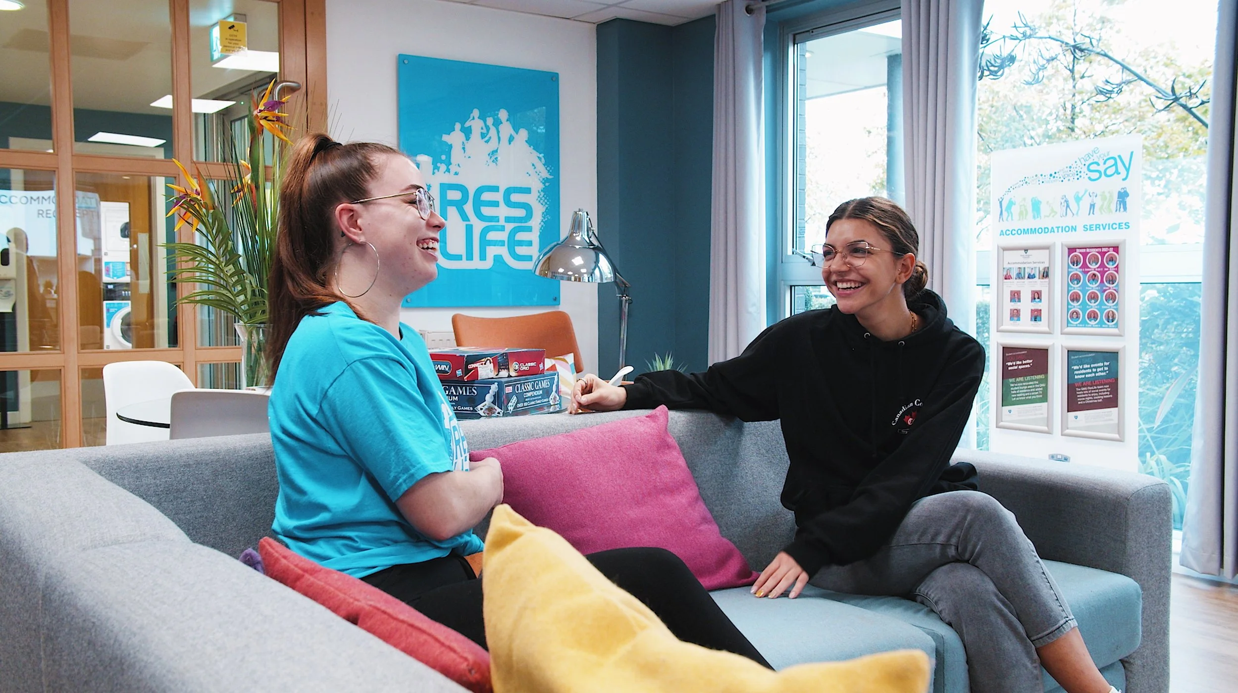 Two young women sitting on a gray couch, engaged in a conversation and smiling. One is wearing a blue t-shirt with glasses, and the other is in a black hoodie with glasses. There are colorful pillows on the couch, and a large window with curtains is behind them. In the background, there are indoor plants, posters on the walls, and signage for accommodation services.