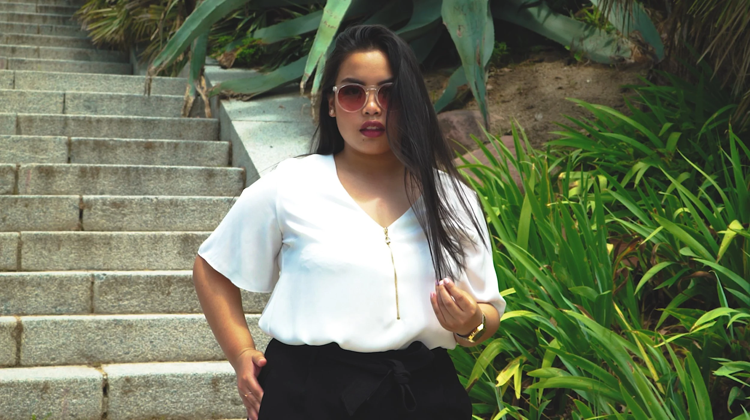 Woman with long hair wearing sunglasses, white blouse, and black pants, standing beside green plants with a stone staircase in the background.