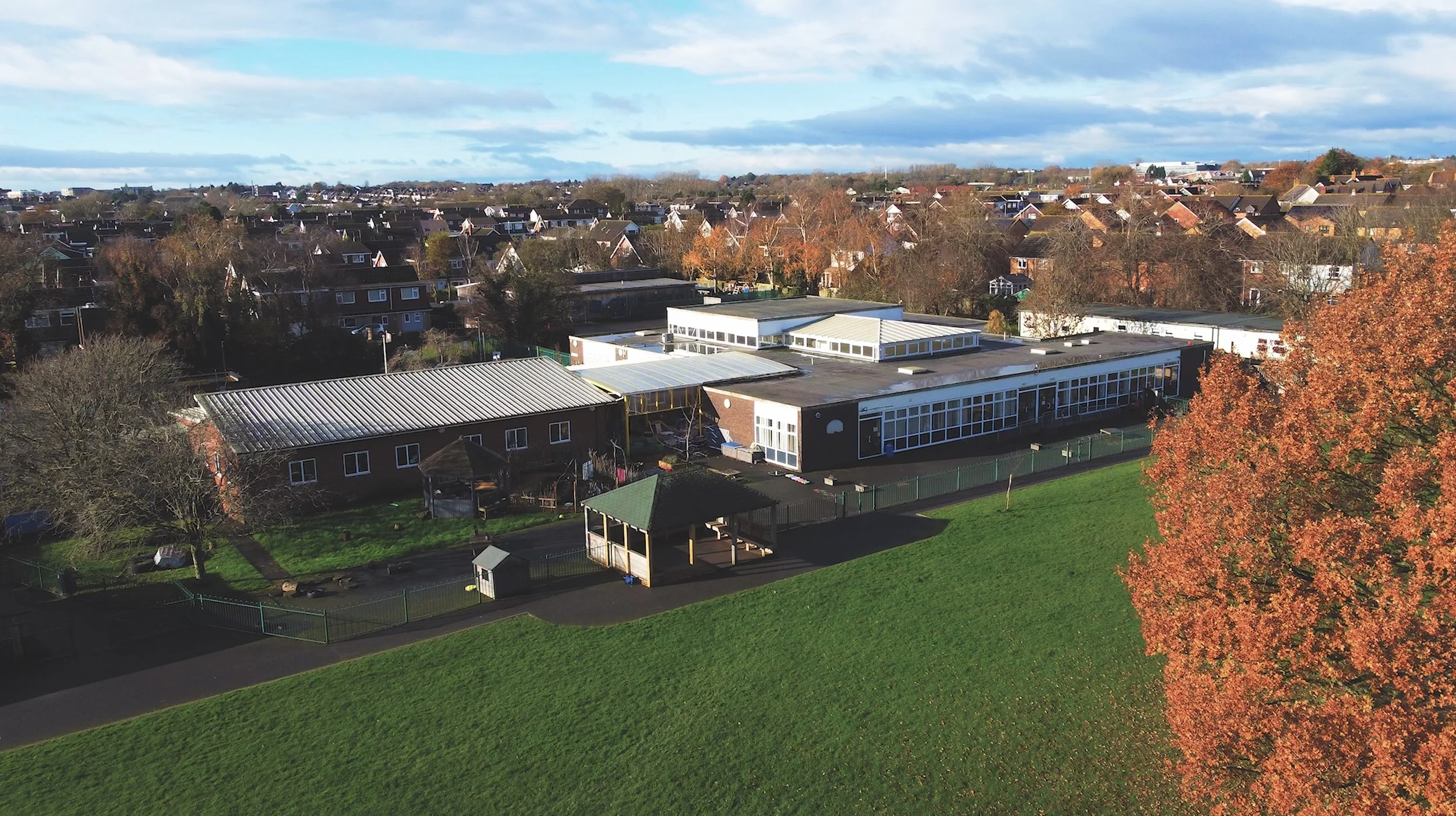 An aerial view of a school with multiple buildings, surrounded by a playground, trees, and a residential neighborhood in the background, under a partly cloudy sky.