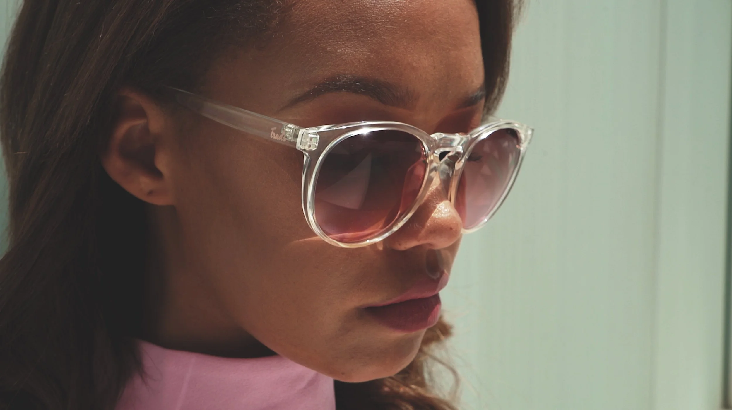 A woman wearing large, clear sunglasses with pink-tinted lenses, looking downwards.