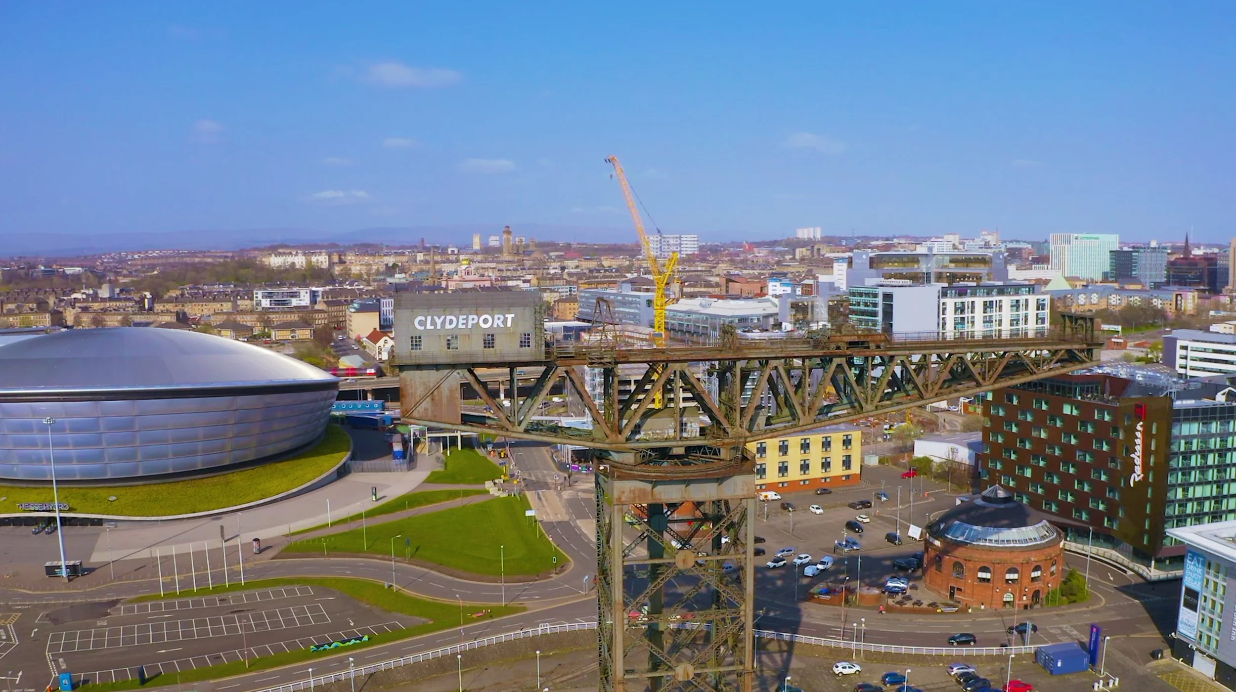 Aerial view of Clydeport with a large, round, silver building on the left, a rusted industrial crane in the center, a modern cityscape with various buildings, and a parking lot, under a clear blue sky.