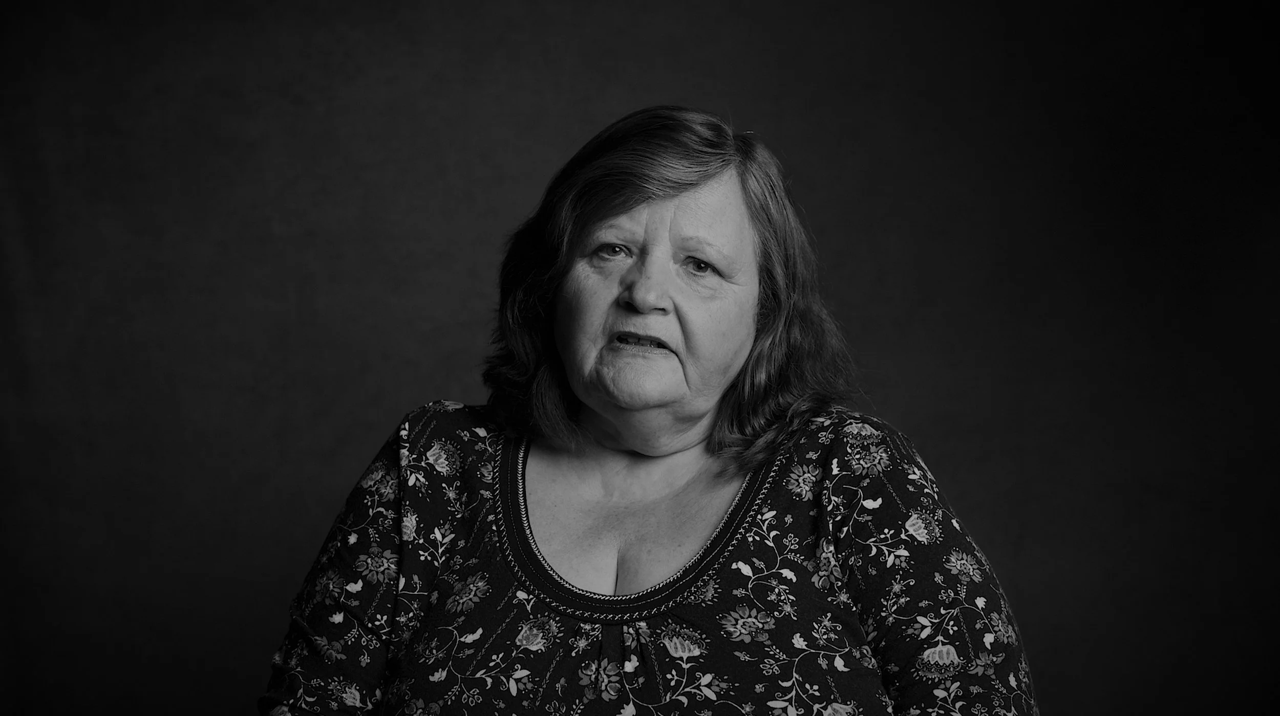 Black and white portrait of a middle-aged woman with shoulder-length hair, wearing a floral patterned top, looking slightly distressed or confused, against a dark background.