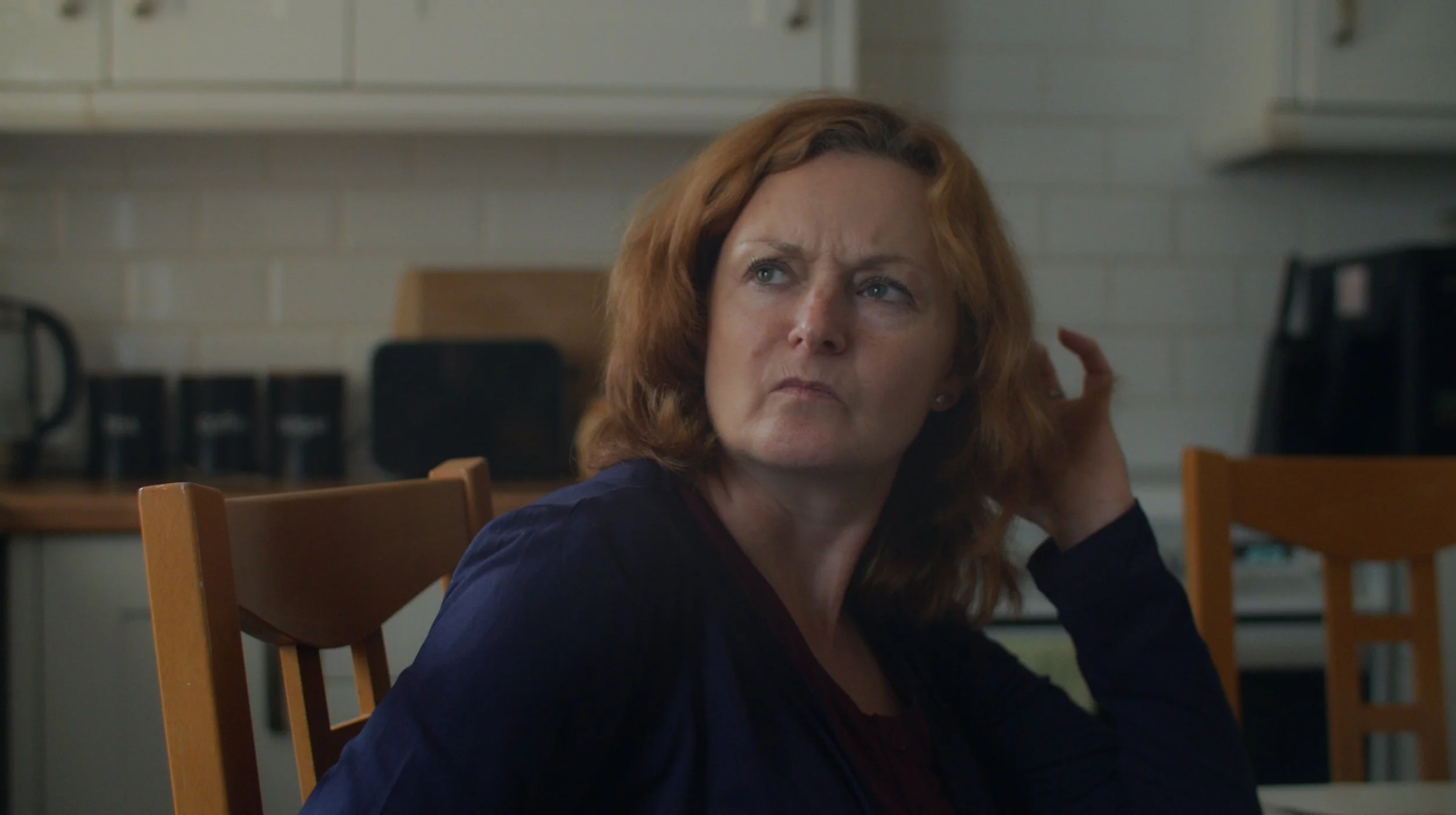A woman with reddish-brown hair sits at a table in a kitchen, looking puzzled or suspicious.